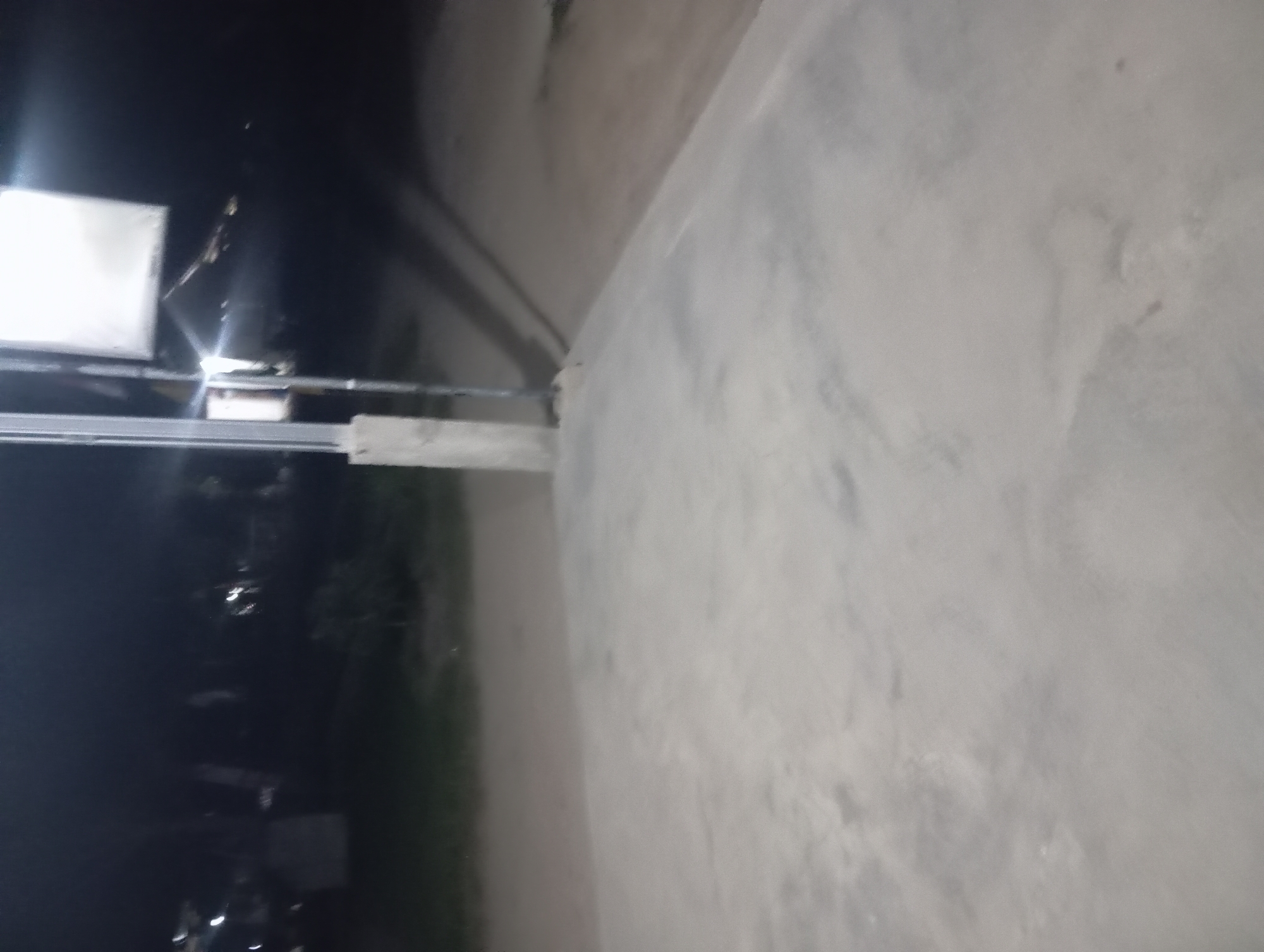 The image captures an outdoor scene at night, likely in Rejosari, Indonesia, with a concrete ground dominating the foreground. A tall, gray pole with a white rectangular sign attached to it stands in the mid-ground, casting a shadow onto the ground behind it. The background is dark and out of focus, with faint hints of vegetation and what might be structures or lights. The overall impression is of a dimly lit, somewhat deserted area, possibly a public space or the exterior of a building.