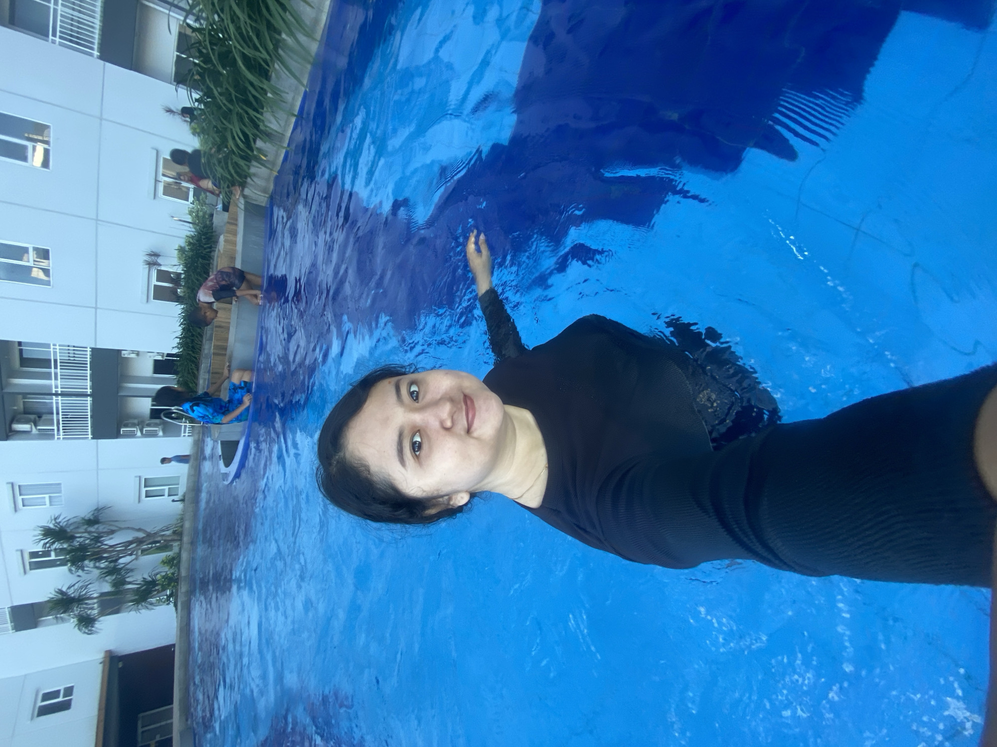 A woman in a black long-sleeved swimsuit is taking a selfie in a blue swimming pool, likely at an apartment complex in Sidoarjo, Indonesia. Several other people are standing at the edge of the pool. A multi-story building is visible in the background.