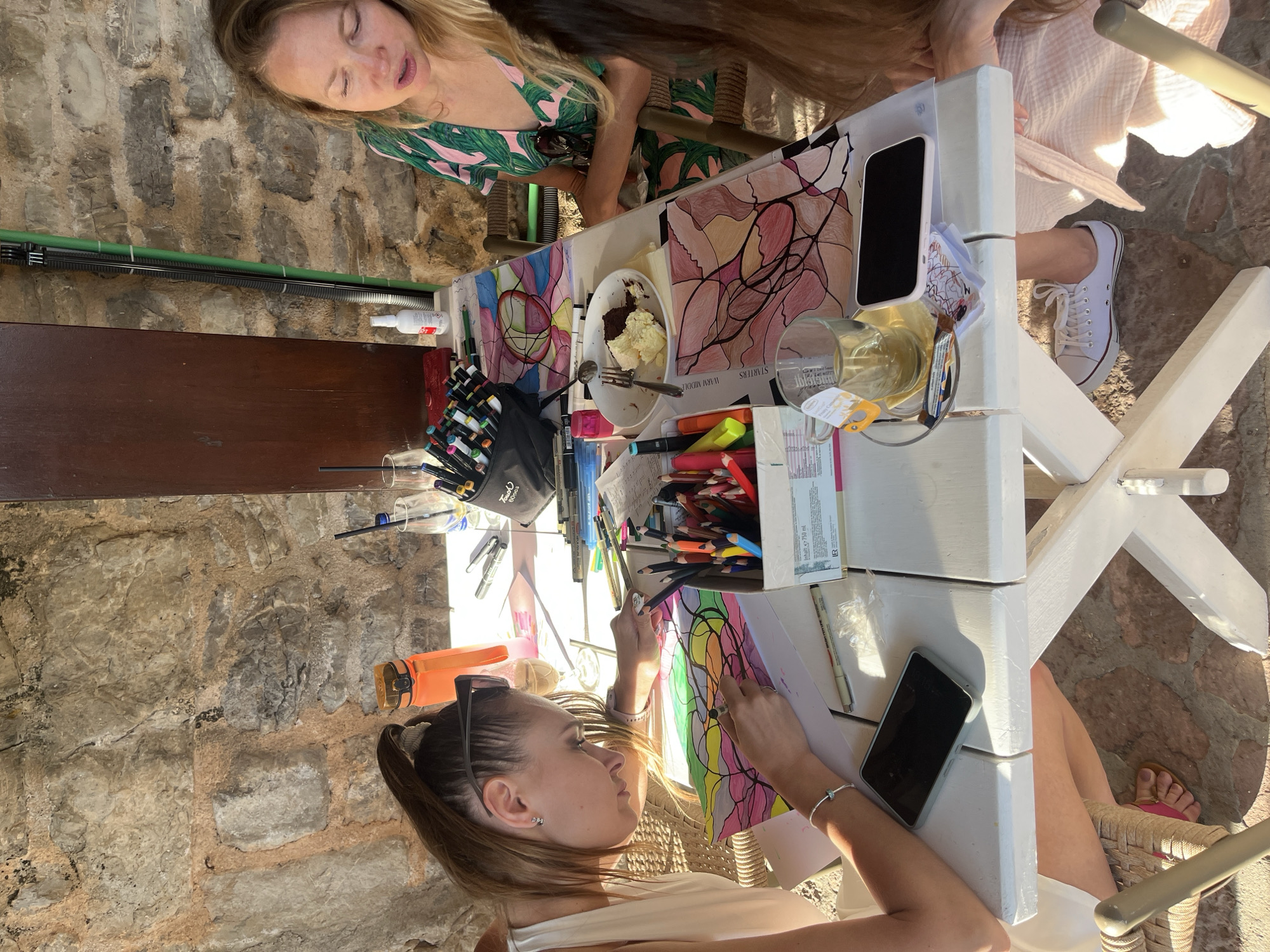 A sunny, outdoor scene depicts three women seated around a white, wooden table with a colorful drawing surface. One woman with her hair pulled back is focused on coloring a drawing with colored pencils, while another woman with blonde hair and a green patterned dress is sitting and the third woman with long brown hair is partially in frame. The table is cluttered with art supplies, including markers, pencils, erasers, and paper. There are also personal items such as two smartphones, a cup of coffee, and a dessert plate with remnants of cake and fork. The setting appears to be an outdoor courtyard with stone walls. The location is in Budva, Montenegro.