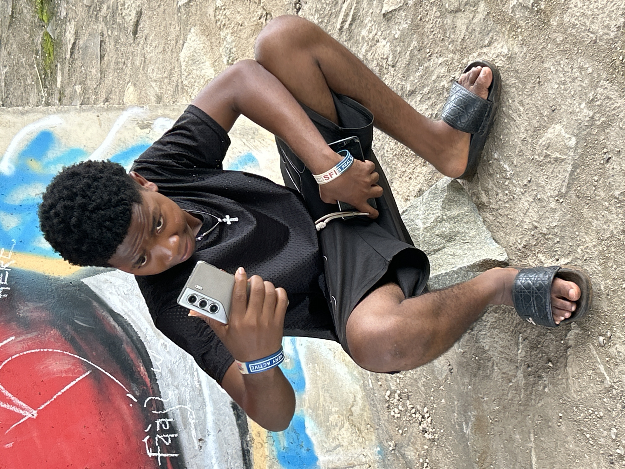 A young Black man is depicted in a rotated outdoor image, appearing to recline against a textured wall or sloped ground in Ihiagwa, Nigeria. He has short, curly black hair and is dressed in a black short-sleeved top, black shorts, and black slide sandals. A silver-colored cross necklace hangs from his neck.
He is holding a light-colored smartphone, featuring a distinct triple-lens camera setup on the back, in his right hand and looking down at its screen. On his left wrist, he wears two bands: a blue one with white text that reads "SFI IS REAL," and a white one with blue text partially visible, saying "VERY ACTIVE."
The background consists of a rough, light-colored concrete or stone wall, showing some signs of moss or discoloration. The ground where he rests is concrete, heavily adorned with colorful graffiti in shades of red, blue, black, and white, with some faint white script visible on the red section. The scene is brightly lit, indicating daytime and clear weather conditions. The man's expression is neutral as he engages with his phone, suggesting a moment of casual activity or relaxation in a public or semi-public outdoor space.
