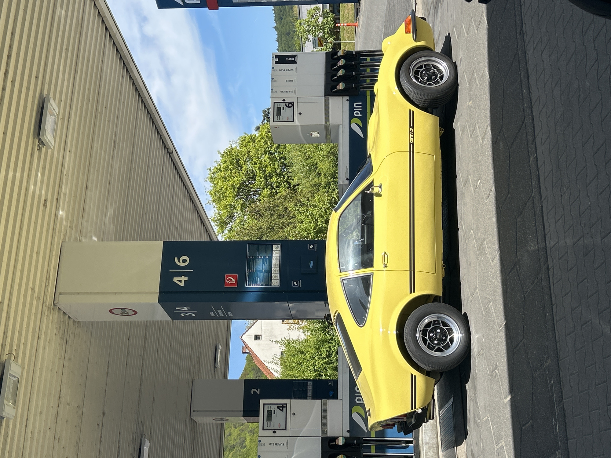 The image shows a yellow Opel GT/J parked at a gas station. The gas station has pumps numbered 2, 3/4, and 4/6. The gas station sells Super E10, Super, and Diesel fuel. The car has black racing stripes and is parked on a paved surface. The sky is blue with some clouds. The location appears to be in a suburban area with trees and buildings visible in the background.