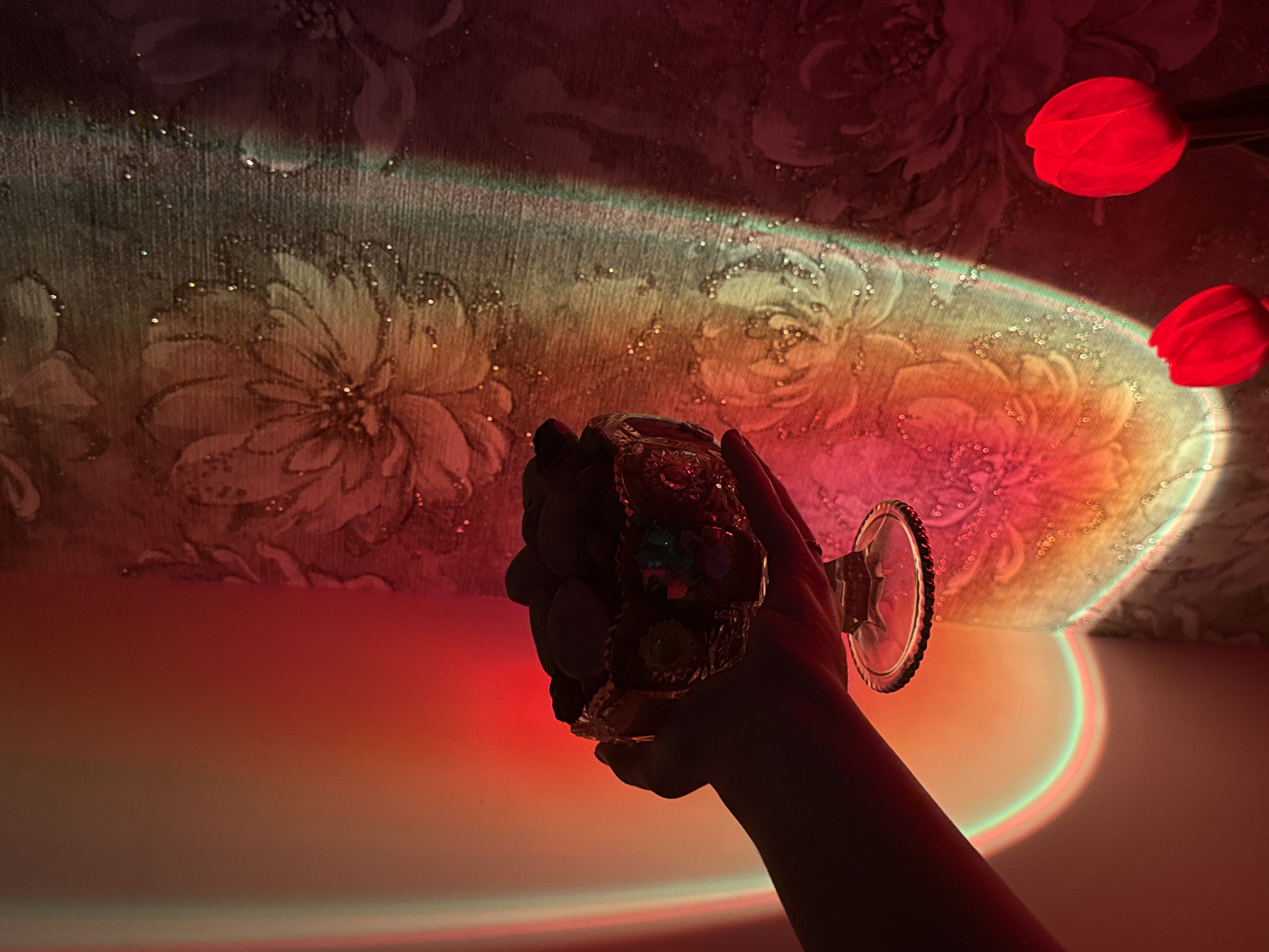 A hand holds a crystal bowl filled with light-colored candies. The bowl is ornate with decorative patterns and colored accents. The background features a patterned wallpaper with floral motifs. Dramatic lighting casts colorful streaks of red, orange, yellow, green, and purple across the scene, creating a vibrant and ethereal atmosphere. Two red tulips are partially visible in the lower right corner, illuminated by the same lighting. The image appears to be an indoor still life, with the location being unknown.