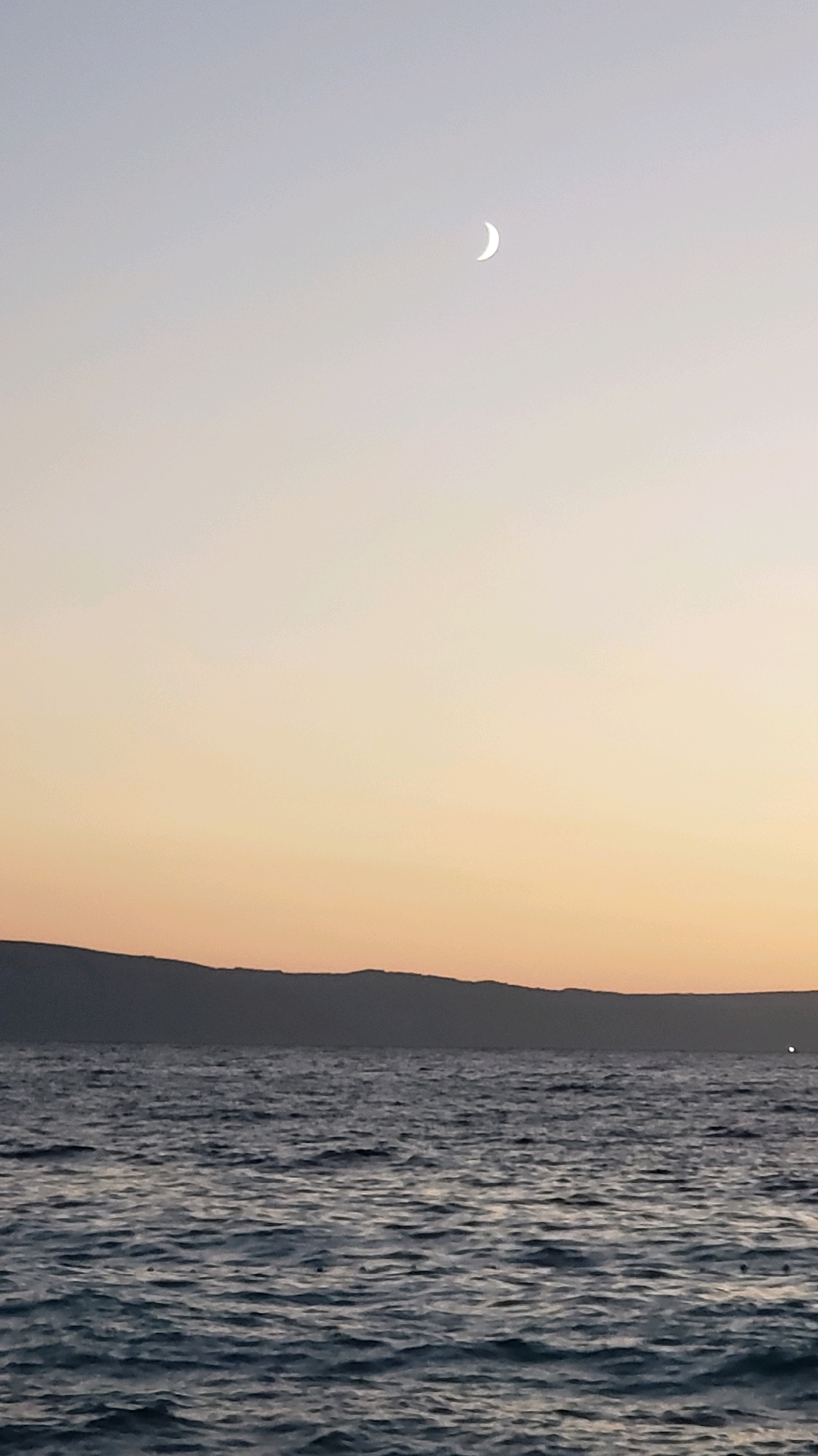 A crescent moon is visible in the sky above a body of water and a silhouetted landmass. The sky transitions from a pale blue at the top to a warm orange-yellow near the horizon. The water is dark blue with visible ripples. The landmass, likely a coastal mountain range in Općina Tučepi, Croatia, is dark and indistinct against the sky. The scene appears to be a coastal landscape at dusk or dawn.
