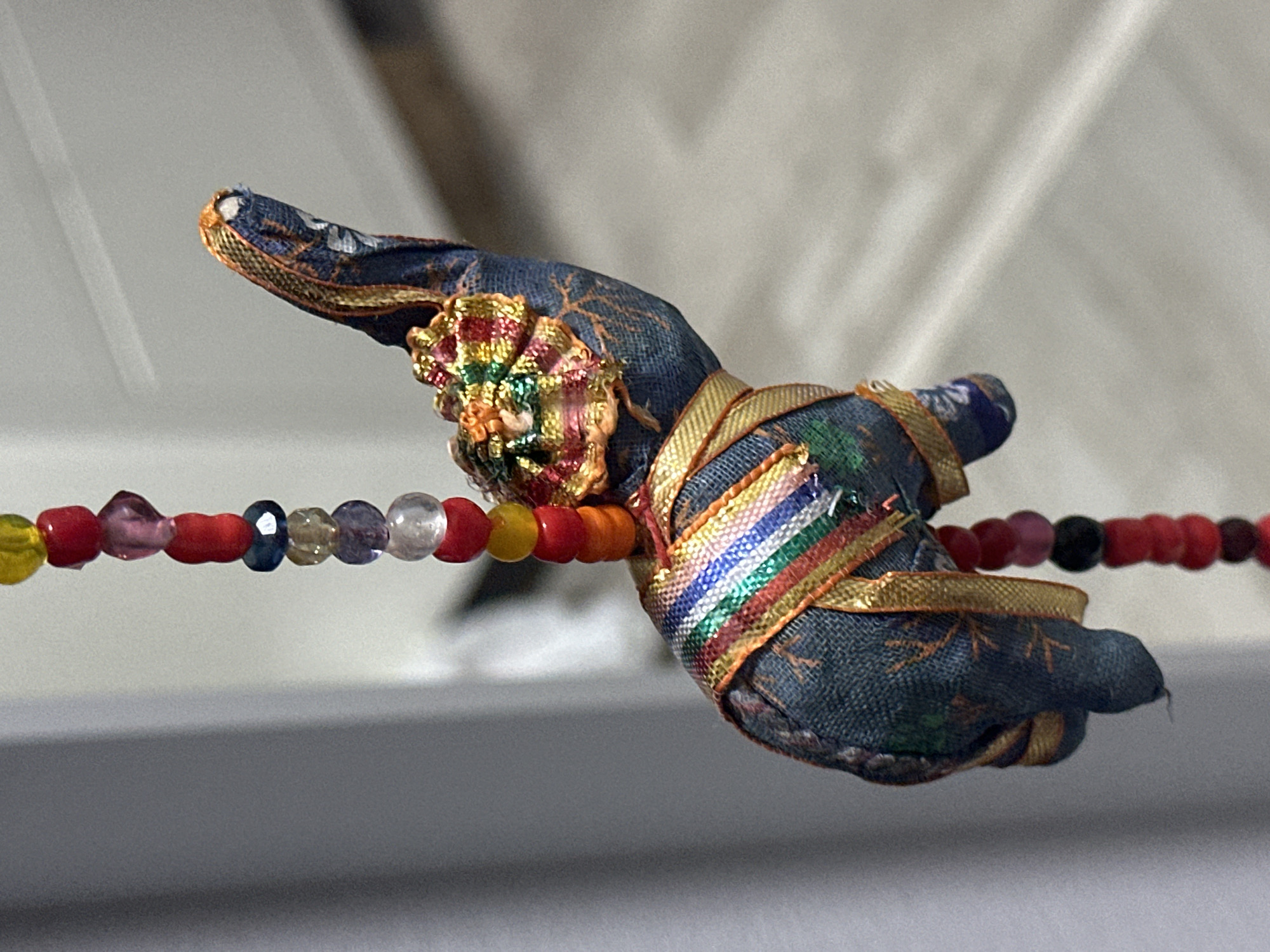A close-up, eye-level shot shows a small, fabric elephant ornament suspended by a string of colorful beads. The elephant is primarily dark blue with a patterned fabric and adorned with gold ribbon and a floral embellishment on its head. The beads are various shapes and colors, including red, yellow, blue, pink, and clear. The background is out of focus, appearing as a softly lit, light-colored surface, possibly a wall or fabric. The lighting is soft, highlighting the textures and colors of the ornament. There is no discernible location in Nevinnomyssk, Russia.