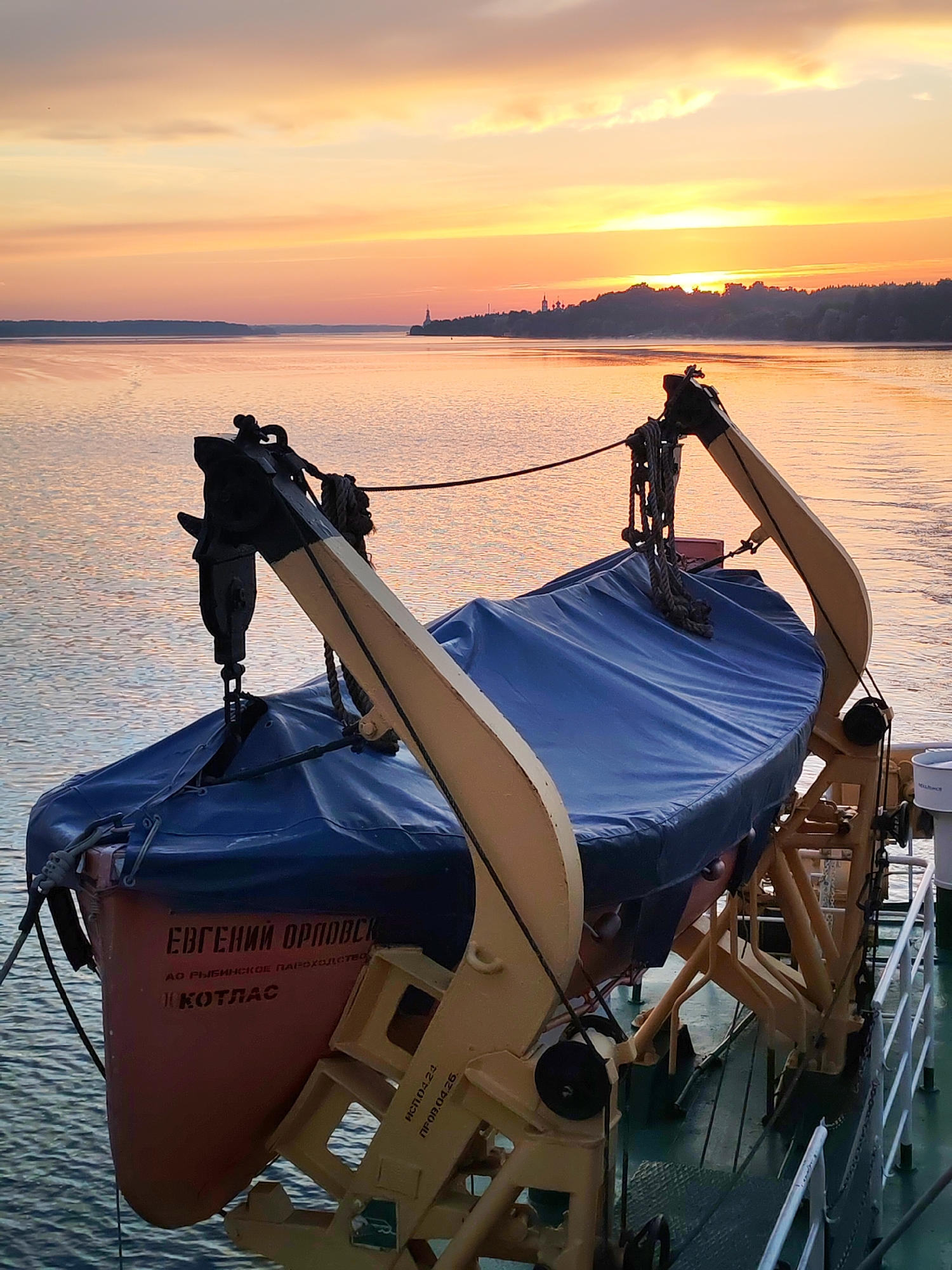 The image shows a life raft secured on the deck of a vessel at sunrise. The sky is filled with hues of orange, yellow, and pink, reflecting on the calm water. The life raft is covered with a blue tarp and is held by a davit system painted yellow and black. In the background, a distant shoreline with trees and a building is visible. Text on the orange hull of the life raft appears to be in Cyrillic. The location is identified as Terekhovo, Russia.
