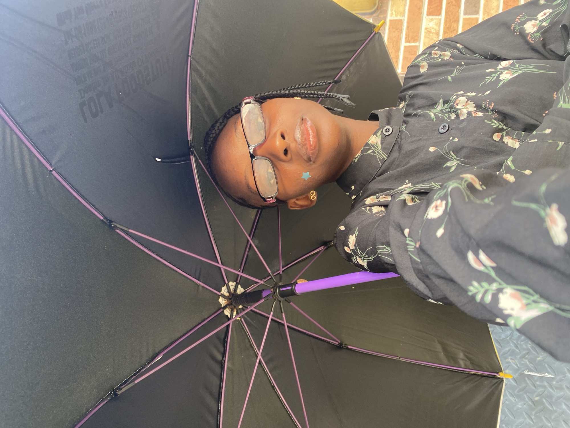 A low-angle, close-up photograph captures a young individual with dark skin, wearing glasses with reddish-purple frames, and braided hair. A small, light blue star-shaped sticker is visible on their right cheek, and they wear gold-colored infinity symbol earrings. They are dressed in a black, long-sleeved, collared shirt with a floral pattern featuring light-colored blossoms and green foliage. The individual holds a black umbrella with purple ribs and a purple handle open above their head. Faint, inverted white text is visible on the inner surface of the umbrella canopy. A light brown brick wall forms part of the background, while a gray, diamond-plate textured surface is visible below the umbrella. The specific location within Iranla, Nigeria, cannot be determined from the visual content.