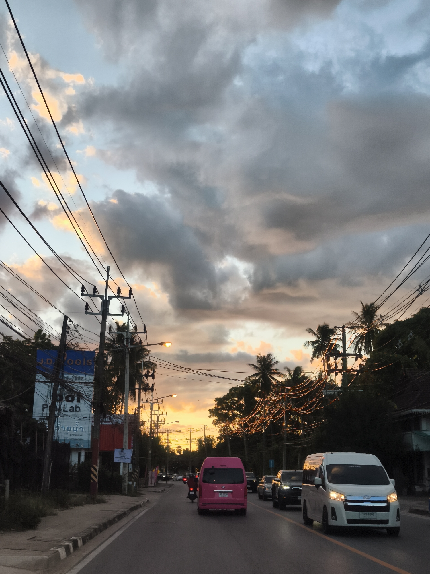 This media file depicts a street scene in Baan Bo Phut, Thailand, during what appears to be sunset or dusk. The sky is dramatic, with a mix of dark, moody clouds interspersed with patches of warm, golden light, suggesting the sun is low on the horizon. The scene is dominated by a road with several vehicles, including a white van, a pink van, and a motorcycle, suggesting moderate traffic.
On the left side of the road, there are utility poles with a complex network of wires, common in many Southeast Asian countries. A tall blue sign with white lettering reads "J.D. Pools" and other unreadable text, likely advertising a business. Further down the street, palm trees line the road, characteristic of tropical locations. String lights are visible, strung between poles and trees, adding a festive atmosphere, especially as dusk settles. The overall mood is somewhat tranquil, with the vibrant sky providing a visually striking backdrop to the everyday activity on the street. The time of day is clearly indicated by the warm, diffused light and the darkening sky. The weather appears to be transitioning, with clouds suggesting a possibility of rain, though the golden hues indicate the sun is still present.