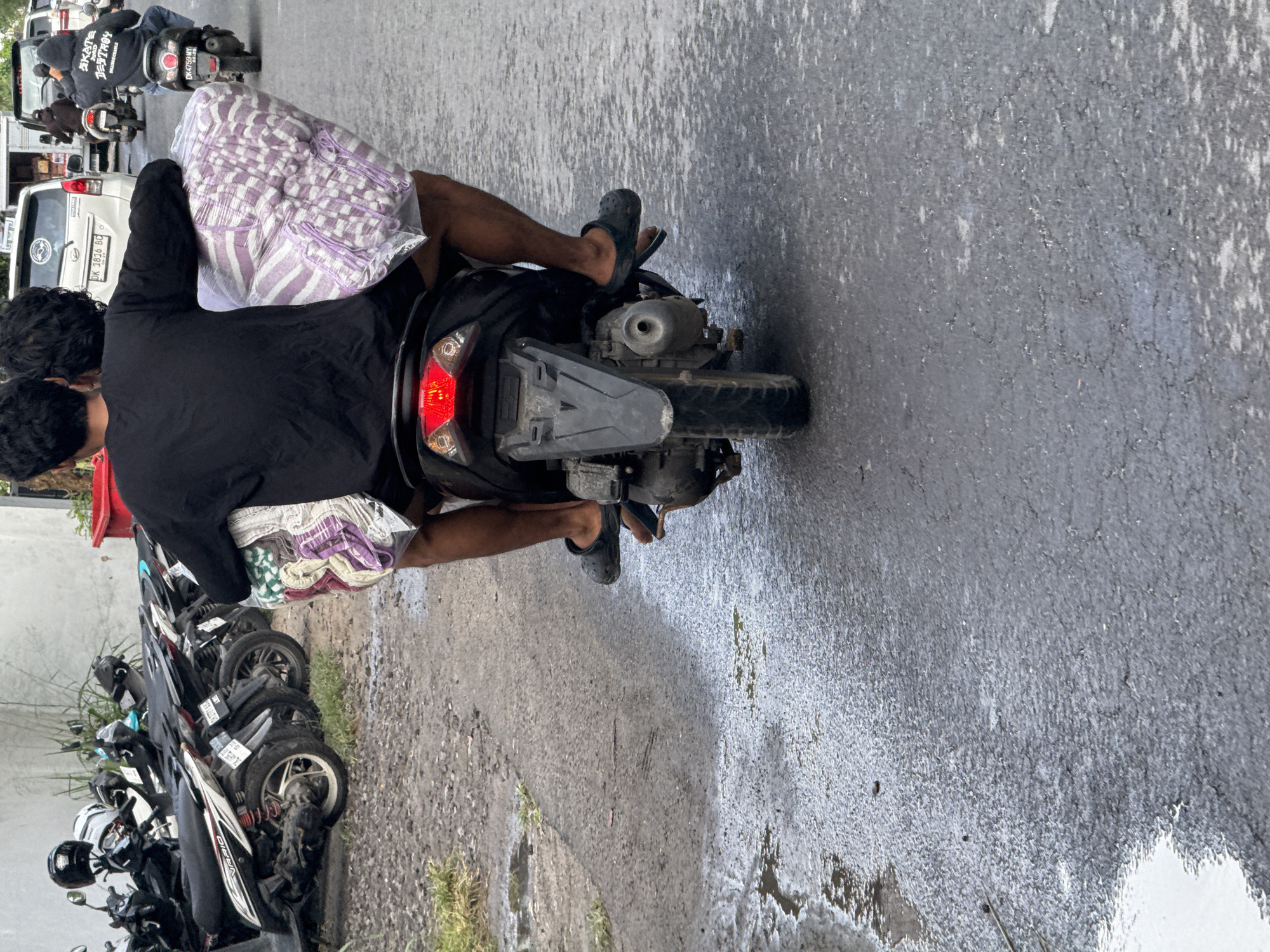A high-angle, sideways perspective shows a person riding a motorcycle on a road in Nusa Penida, Indonesia. The rider wears a black T-shirt, striped shorts, and black sandals. A package is between the driver and the back seat passenger. The road is wet and gray, likely from recent rain. In the background, a line of parked motorcycles is visible, along with a white van and other vehicles. Another motorcyclist is ahead of the subject. The setting appears to be an urban or semi-urban street.