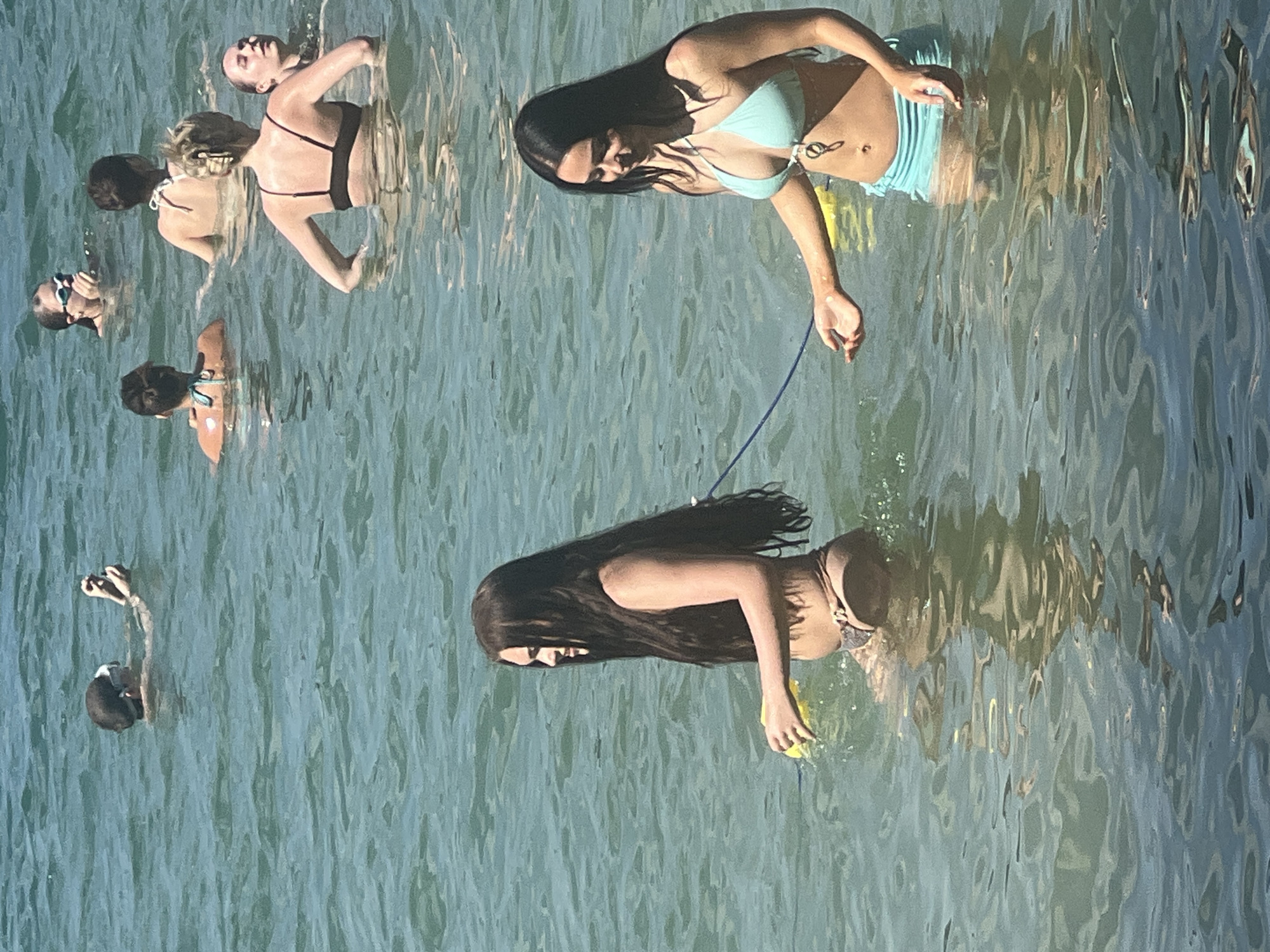 The image shows a group of people in the water. The subjects appear to be women wearing swimsuits. The scene is set in a body of water, likely a lake or the sea, with clear, rippling blue-green water. Several individuals are partially submerged, with some swimming and others wading.
In the foreground, a woman with long dark hair is swimming. She is wearing a bikini and appears to be holding a yellow object, possibly a buoyancy aid or toy. Her expression is not clearly visible. Behind her, and slightly to the left, another woman is visible, also in the water. Further to the left and behind them, a group of three other individuals are swimming or floating. One of them is wearing a black bikini, another a lighter colored bikini, and a third has blonde hair. The image is taken from a high angle, looking down onto the water.
The lighting suggests it is daytime, likely sunny, given the reflections on the water. The water's surface is disturbed by ripples, indicating some movement. The overall impression is one of leisure and recreation in a natural water setting. There is no visible text in the image. The location is Vilnius, Lithuania.