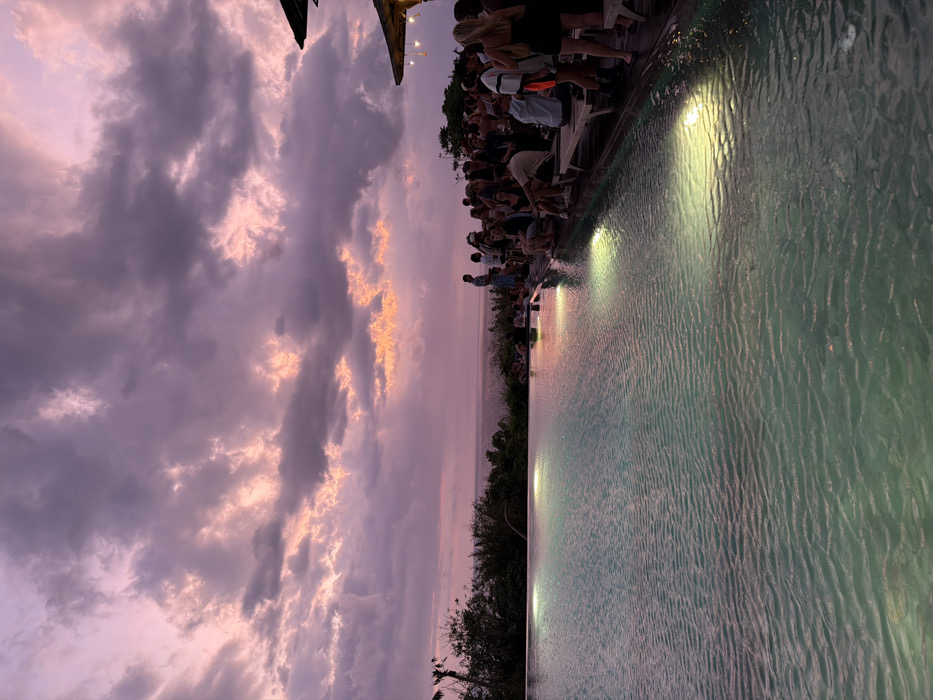 The image depicts an outdoor evening scene at an infinity pool facility in Uluwatu, Indonesia, during twilight hours. The sky above is characterized by an extensive cloud cover with pronounced purple and pink hues, indicative of a sunset, interspersed with darker grey cloud formations. The horizon line separates the sky from a calm, dark-blue ocean visible in the mid-ground.
In the foreground, a large, light-blue infinity pool occupies the lower left portion of the frame, showing ripples on its surface. Four submerged lights within the pool emit distinct yellow-green glows, illuminating the water. To the immediate right of the pool, a wooden deck extends, accommodating a gathering of approximately 30-40 individuals. These individuals are predominantly standing in various groups, engaged in social interactions, while some are seated on wooden lounge chairs or benches. A few individuals are visible further back, closer to the ocean's edge, also gathered socially. One person stands near the far edge of the infinity pool, facing the ocean. On the far right, a portion of a building or canopy roof, constructed from a natural material and adorned with string lights, is visible. Lush green vegetation borders the scene on the left, obscuring part of the ocean view.