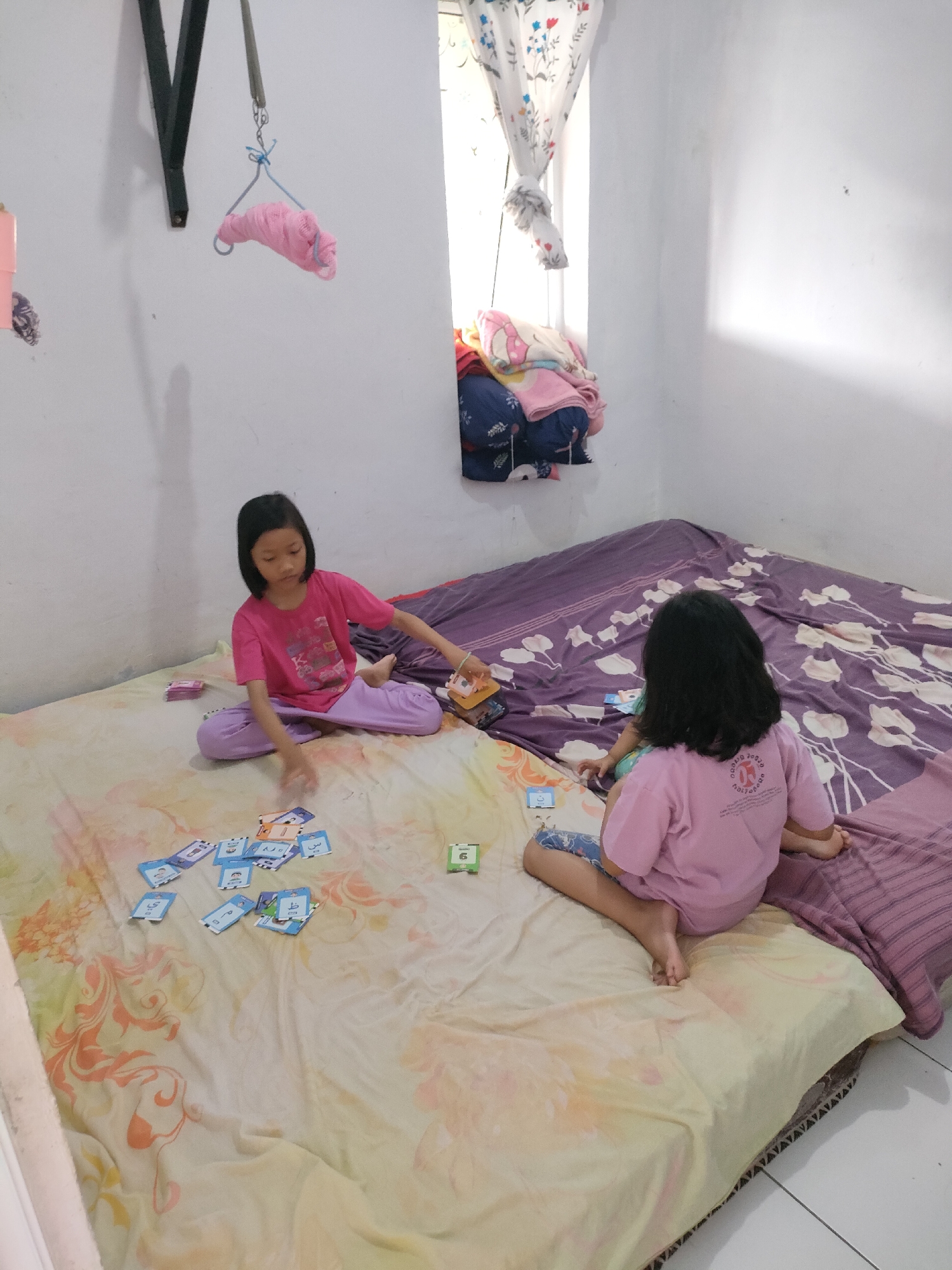 Two young girls, one in a pink t-shirt and purple pants, and another in a pink t-shirt and blue patterned shorts, are playing with cards on a bed. The girl in the pink t-shirt and purple pants is sitting closer to the camera, with a pile of cards spread out in front of her. The other girl is sitting further back, facing away from the camera. The bed is covered with a floral patterned sheet. In the background, there is a window with a curtain, and a pile of clothes. The room appears to be a bedroom, possibly in a residential area of Unknown, Indonesia.