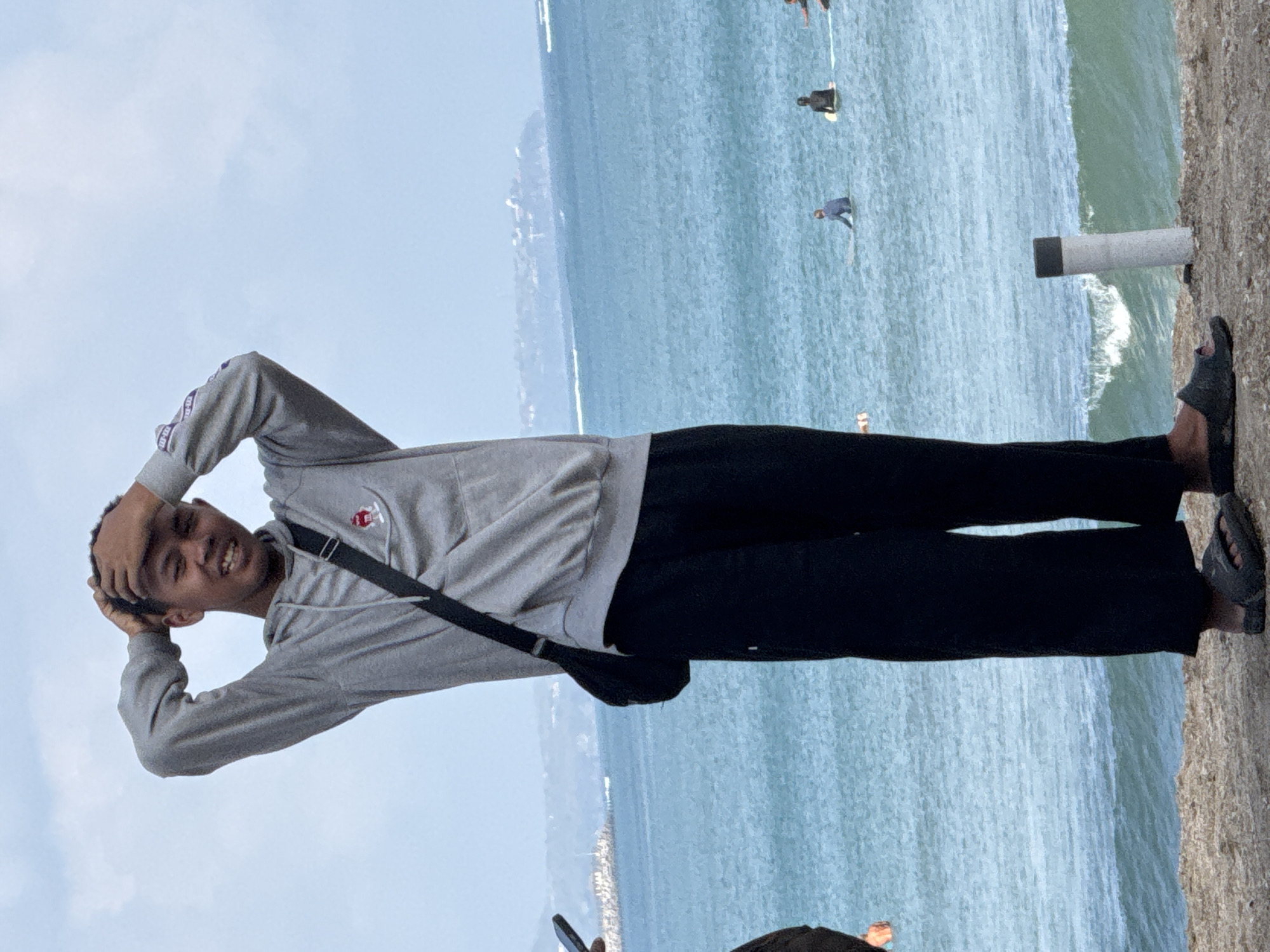 A full shot shows a young man standing on a beach in Kuta, Indonesia. He is wearing a grey hooded sweatshirt and black pants, with a black bag slung across his chest. His hands are placed on his head, and he is smiling. The ocean is in the background, with several surfers in the water. The sky is overcast.