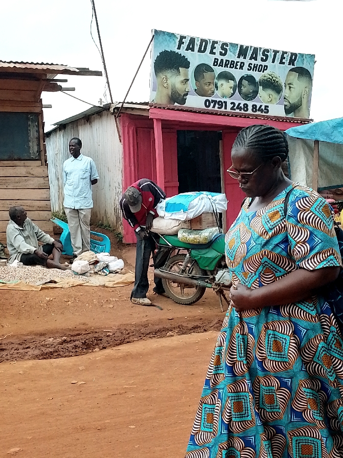 The image shows a street scene in Lwandeti, Kenya (latitude: 0.563, longitude: 34.806), with a woman in the foreground, a barber shop sign in the background, and several men in between. The woman is wearing a blue and gold patterned dress, and she is standing on a dirt road. Behind her, to the left, a man in a blue shirt is standing. He appears to be looking at something off-screen, out of frame. Behind him, to the left, is a man seated on the ground, holding a white object in his lap. To the right of the woman, a man wearing a red shirt and black pants is standing near a motorcycle. The motorcycle has a blue tarpaulin, and is being loaded with other bags. There are bags on the ground near the motorcycle, and a few other items, but it is not clear what they are. The man in the red shirt appears to be putting something into a bag, and the motorcycle appears to be parked, as the rider has stepped off of it. There is a sign above the motorcycle that says "Fades Master Barber Shop" with a phone number underneath. The sign has several pictures of men with different hairstyles. It is unclear what the man in the red shirt is doing. The woman is facing towards the camera, while the three men behind her are all facing to the left. There is a wooden structure behind the men, which may be part of the barber shop. The street is a dirt road, with a muddy patch near the motorcycle. It is a sunny day.