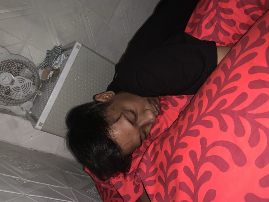 A person sleeps on a red pillow with a coral design. They are wearing a black top. Beside them is a white woven plastic storage unit. A small white fan sits on top of the unit, with some items placed behind it. The floor is tiled in a white geometric pattern. The picture has a slightly rotated perspective.