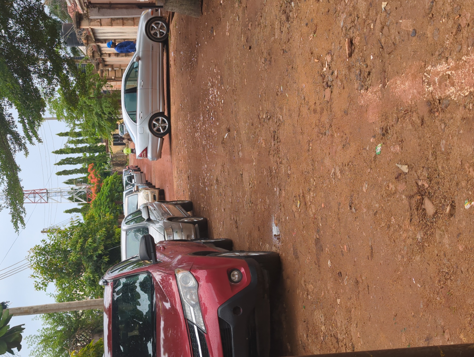 The image displays a line of cars parked on the side of a reddish-brown dirt road in what appears to be a residential or semi-rural area, consistent with Unguwar Rimi, Nigeria.
The closest vehicle is a red SUV or crossover, with visible raindrops on its surface, indicating recent rain. Behind it, a silver sedan, identifiable as a Mercedes-Benz C-Class (W203 generation), is parked. Several other vehicles, mostly light-colored, are visible further down the line.
The road itself is unpaved, made of reddish-brown earth and mud, appearing wet and uneven in several spots, with scattered small stones and debris. To the left of the parked cars, a dense line of green trees and shrubs forms a natural barrier, with a light-colored wall or fence visible behind them. A dark water tank is mounted on a structure further back. In the background, a prominent red and white lattice communications tower stands tall against the sky.
One person wearing a blue shirt and cap is partially visible standing near the silver Mercedes-Benz in the distance. Another individual is faintly discernible near the front of the red car, somewhat obscured by the trees.
The scene takes place during the daytime, likely under overcast conditions or following rainfall, given the wet appearance of the cars and ground. There is no discernible text present in the image.