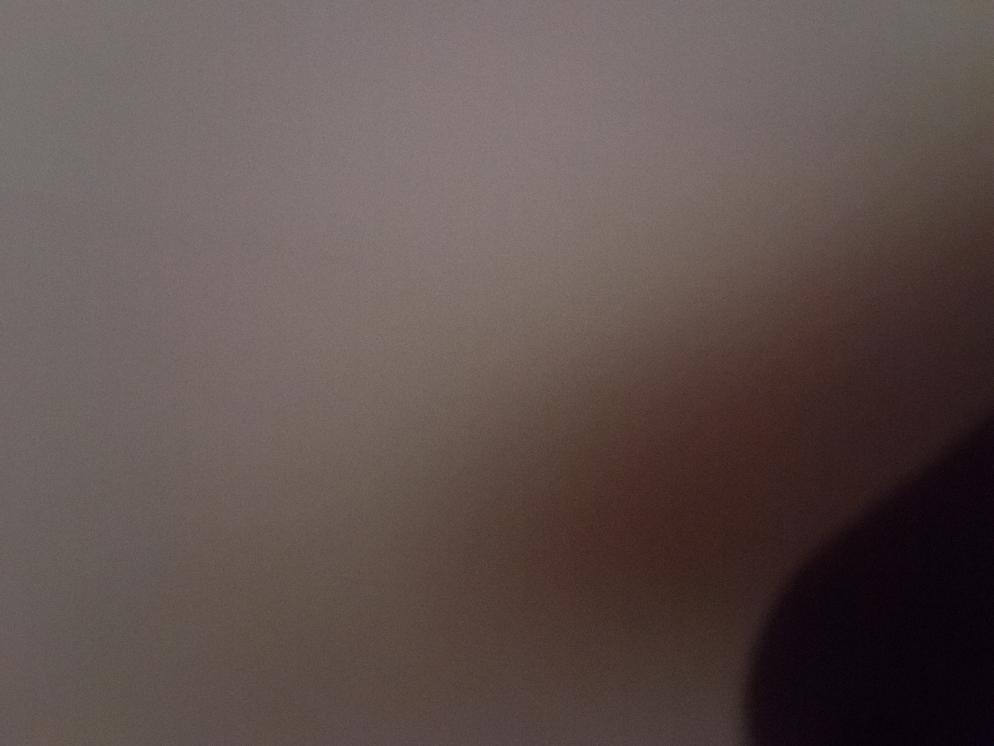 The image is a blurred photograph, appearing to be taken indoors. The foreground is dominated by a dark, amorphous shadow that occupies the lower left portion of the frame. The background is a gradient of muted, neutral tones, transitioning from a lighter, grayish-brown in the upper right to a darker, reddish-brown in the lower left. The overall lighting is dim, and the image exhibits a grainy texture. There are no discernible objects, people, or actions visible in the scene. The location in Jauro Yinu, Nigeria, is not evident from the visual information provided.