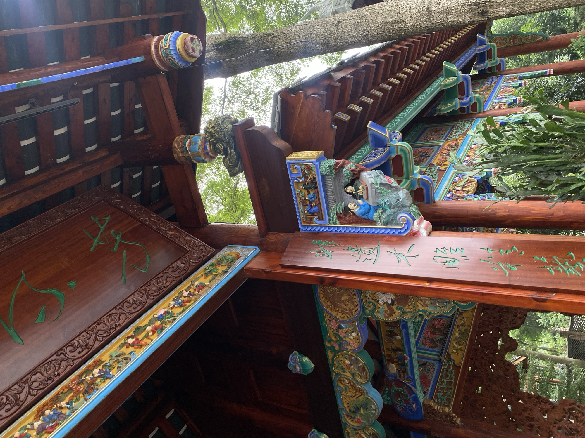 The image shows a detailed view of a traditional Chinese pavilion, likely located in Chongzhou City, China, known for its cultural heritage. The pavilion features intricate wooden carvings and vibrant painted decorations. The roof is made of dark tiles supported by reddish-brown wooden beams and pillars. The eaves are adorned with colorful patterns, including depictions of figures and mythological creatures, rendered in blue, green, gold, and red. Green Chinese calligraphy is inscribed on the wooden surfaces.
In the foreground, the focus is on a large wooden pillar with carved inscriptions and ornate decorative elements. Below the eaves, a row of bracket supports painted in blue and green is visible. A lush green plant, possibly a cactus or succulent, grows in front of the pavilion.
To the right, a large tree trunk with rough bark stands next to the pavilion, its branches extending upwards and partially obscured by the roof. The background reveals more of the pavilion's structure and hints at a natural, possibly garden-like, environment with trees and foliage. The lighting suggests it is daytime, with diffused light filtering through the leaves. The overall impression is one of traditional craftsmanship and artistic expression within a serene setting.