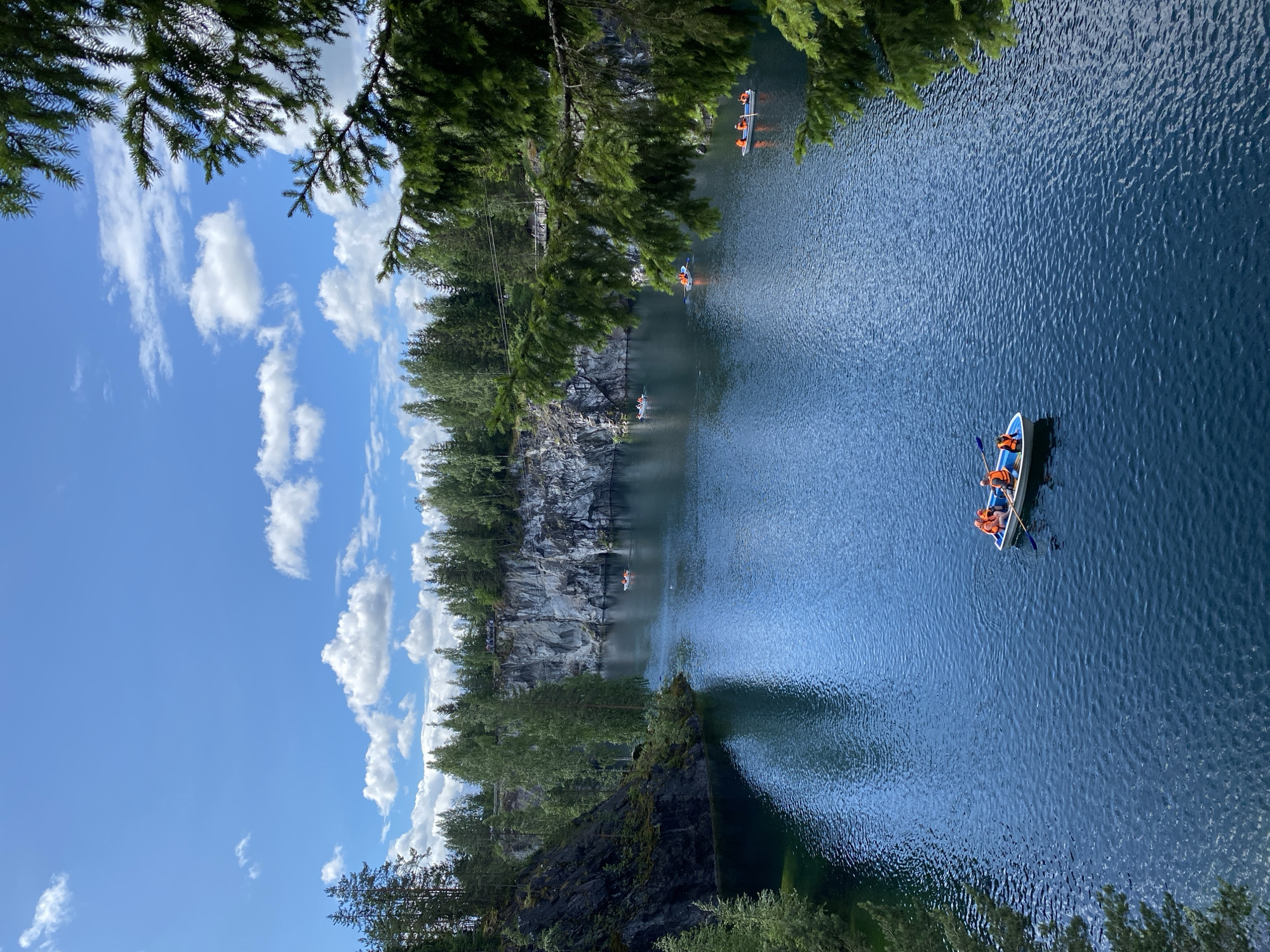 This is a high-angle, full-frame, eye-level shot of Ruskeala Mountain Park in Russia. The scene features a body of tranquil, deep blue water, likely a lake or quarry, surrounded by steep, rocky cliffs. Lush green trees line the top of the cliffs and extend to the horizon. The sky above is a clear, vibrant blue, dotted with scattered white, fluffy clouds.
Several small boats with people wearing orange life vests are visible on the water. In the foreground, closer to the bottom center, a boat carries at least three individuals engaged in rowing or paddling. Further out in the water, other boats are scattered, with some appearing to have fewer occupants.
The rocky cliffs display a rough, textured surface, with some areas showing dark, mossy patches and others appearing lighter in color, possibly due to marble formations. The water is disturbed by the movement of the boats, creating ripples that reflect the sunlight. A few tall evergreen trees are prominently featured in the right foreground, with their branches extending into the frame, partially obscuring the view of the scene. The overall impression is one of natural beauty and recreational activity.