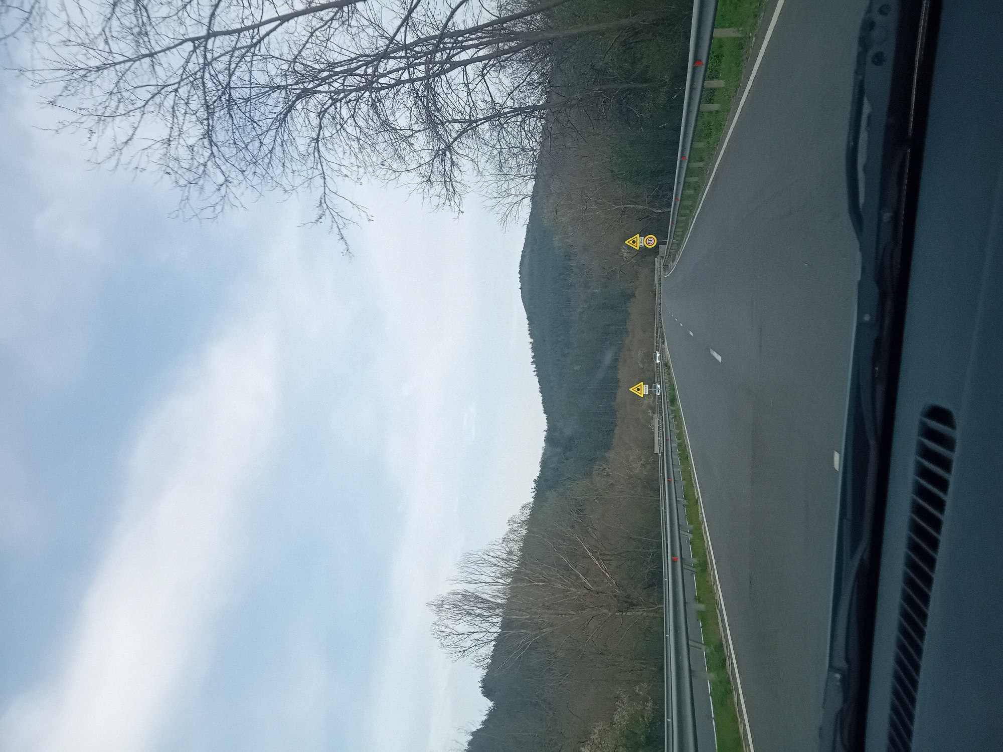 The image depicts a view from inside a vehicle traveling on a road. The road has two lanes in the visible direction with a guardrail on the right side. The sky is cloudy, and there are bare trees and a hillside visible adjacent to the road. There are road signs visible on the side of the road. The specific location and city are unknown.
