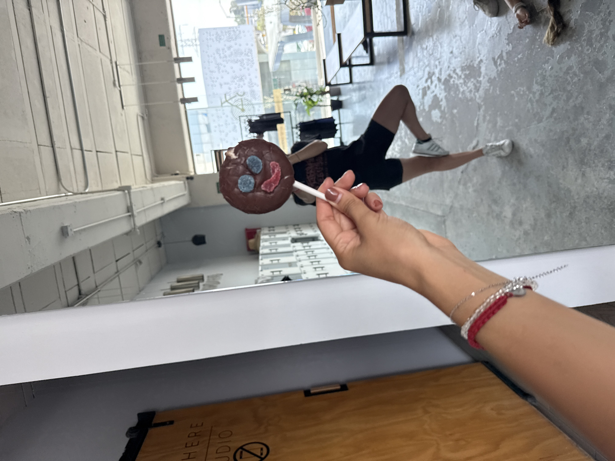 A person is holding a chocolate lollipop with blue and red candy decorations in front of a mirror, seemingly taking a selfie. The reflection in the mirror shows a person lying on the floor in black shorts and white sneakers. The location appears to be inside a building with large windows, potentially a studio or commercial space. In the background, through the windows, there are blurred urban elements suggestive of an outdoor environment. Based on contextual clues like the presence of a sign that says "Studio" on the furniture, the image may be a studio space in Puebla, Mexico. The lighting is predominantly bright, natural light coming through the windows.