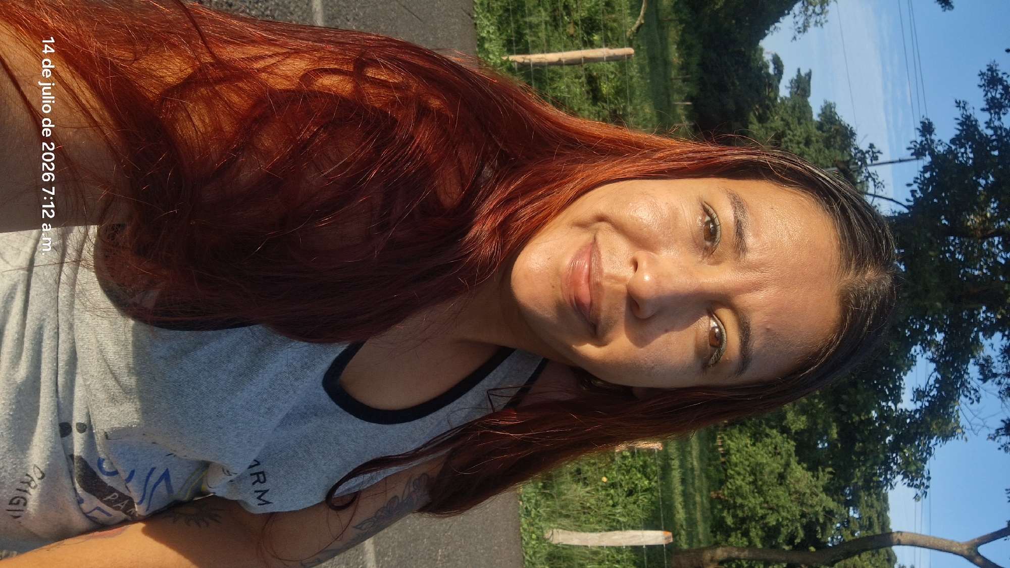 A medium shot shows a woman from the chest up, facing the camera. She has long, reddish-brown hair that cascades over her shoulders and chest. Her skin is tanned, and she is smiling with her lips slightly parted. She is wearing a light gray tank top with black trim around the neckline and armholes. The tank top has some text and designs on it, but they are partially obscured. On her left arm, a tattoo is visible.
The background is a natural outdoor setting. To the left and behind her, there is a grassy area with wooden fence posts. Further back, there are trees and foliage under a clear blue sky with some wispy clouds. To the right, there is a paved surface, likely a road, and more greenery. The lighting suggests it is daytime, possibly morning or late afternoon, given the golden hue on her skin. The date and time stamp in the bottom left corner reads "14 de julio de 2026 7:12 a.m." indicating the image was taken on July 14, 2026, at 7:12 AM. The location is identified as Vallecito, Venezuela.