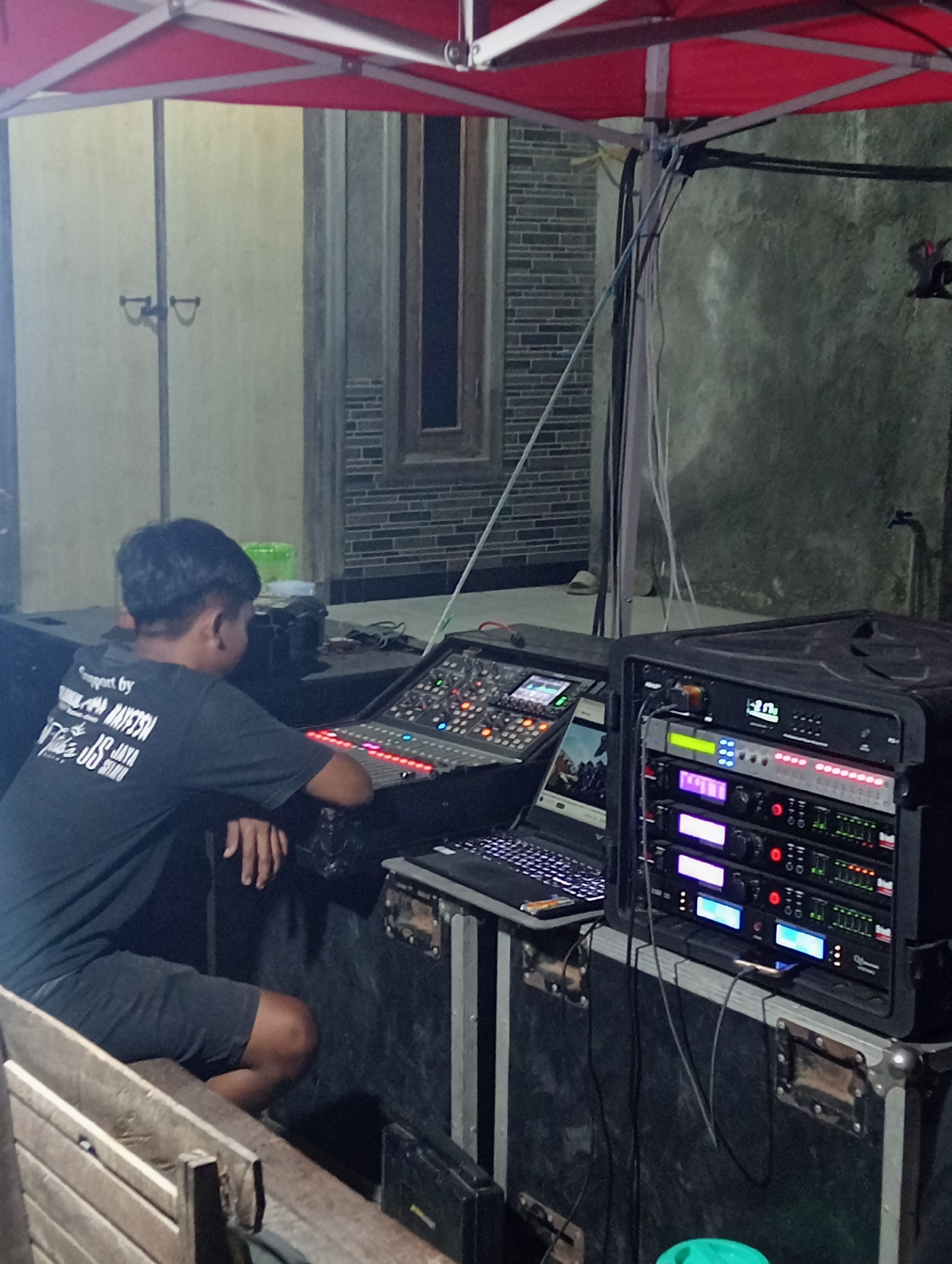 In Tanjunganyar, Indonesia, a young man is seated behind a sound mixing console and audio equipment, likely for an outdoor event. He is wearing a dark t-shirt and shorts. The equipment includes a large mixing board with numerous knobs and faders, a laptop, and several rack-mounted audio processors with illuminated displays. The scene is set up under a red tent, with a brick wall and a window visible in the background, suggesting a residential or public area. Cables are visible, connecting the various components.