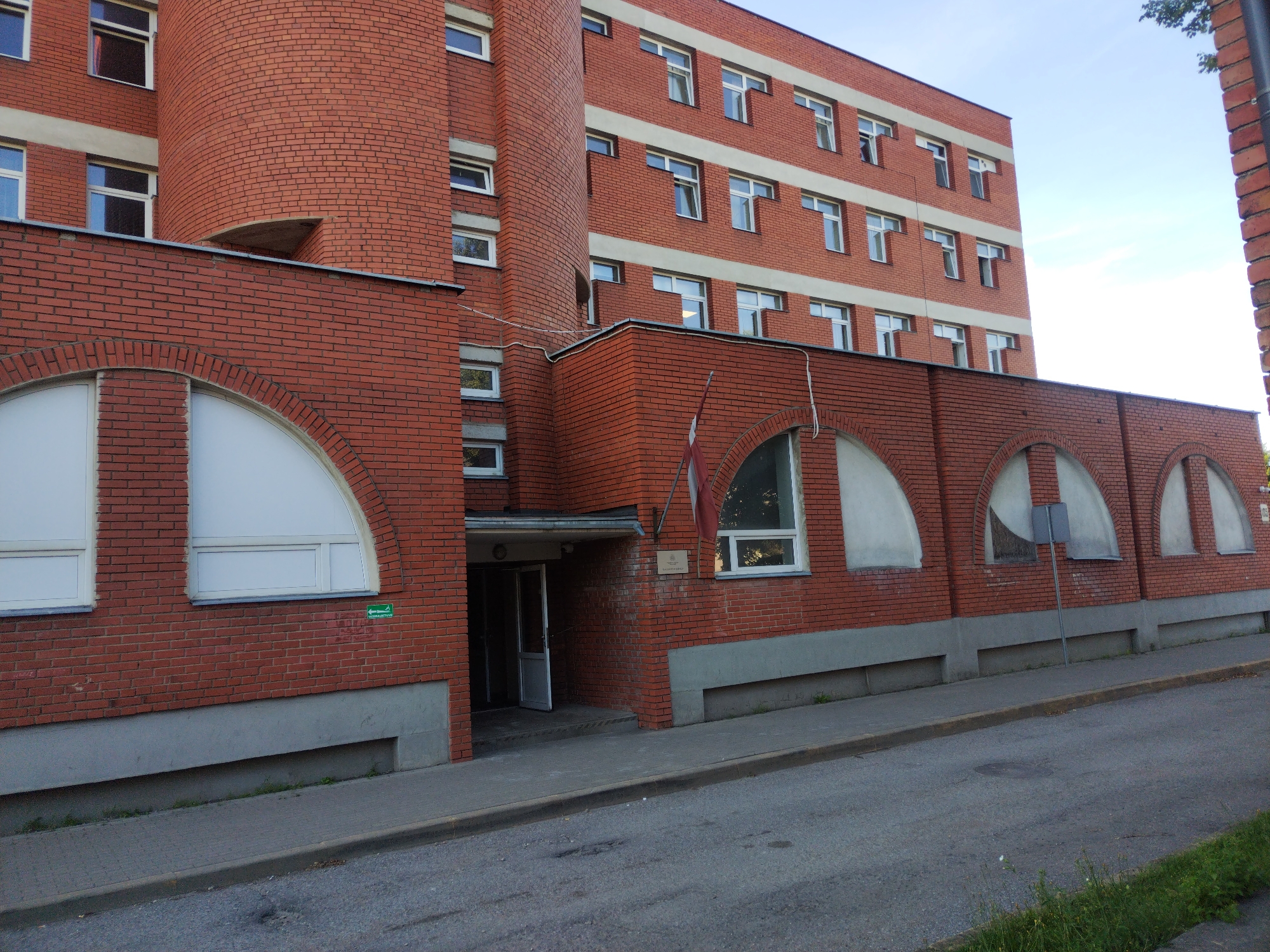 A low-angle, full shot captures the exterior of a brick building in Daugavpils, Latvia, under a clear blue sky with wisps of clouds. The building is predominantly constructed from red brick, with horizontal bands of lighter-colored material, possibly concrete or a lighter brick, accentuating its structure. The main building on the right is a multi-story structure with numerous windows, each featuring white frames and reflective glass, suggesting the windows are closed. The windows are arranged in a repeating pattern across each level.
To the left, a curved section of the building also made of red brick is visible, with a few small, rectangular windows. A lower section of the building in the foreground is also brick, featuring several large arched windows with white opaque panes. A small green sign with an arrow is visible on this section. An entrance with glass doors and a prominent red and white flag, likely the Latvian flag, hanging to the right of the entrance is also visible.
The building is situated along a paved road with a curb and a sidewalk. There is some sparse green vegetation growing along the base of the building and at the edge of the sidewalk. The overall scene depicts an institutional or public building in an urban environment.