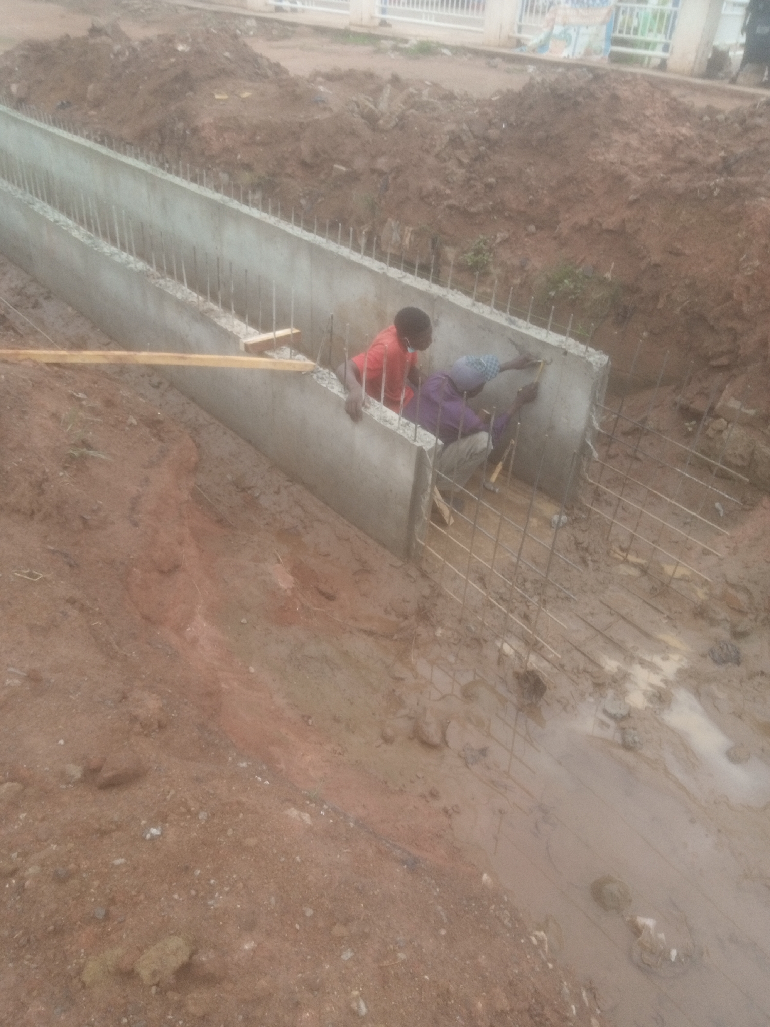 This image depicts an outdoor construction scene in Kakuri Gwari, Nigeria. Two individuals are actively engaged in building a concrete structure, possibly a drainage system or foundation. One person, wearing a red shirt, is standing and appears to be supervising or assisting. The other, dressed in a purple shirt and headscarf, is kneeling and working on the concrete wall, possibly cementing or reinforcing it. The surrounding environment is muddy and disturbed earth, indicating recent excavation or construction activity. Reinforcement bars are visible protruding from the concrete structure and extending into the ground. A wooden plank rests on the edge of the concrete. The overall scene suggests ongoing civil engineering work in a developing area.