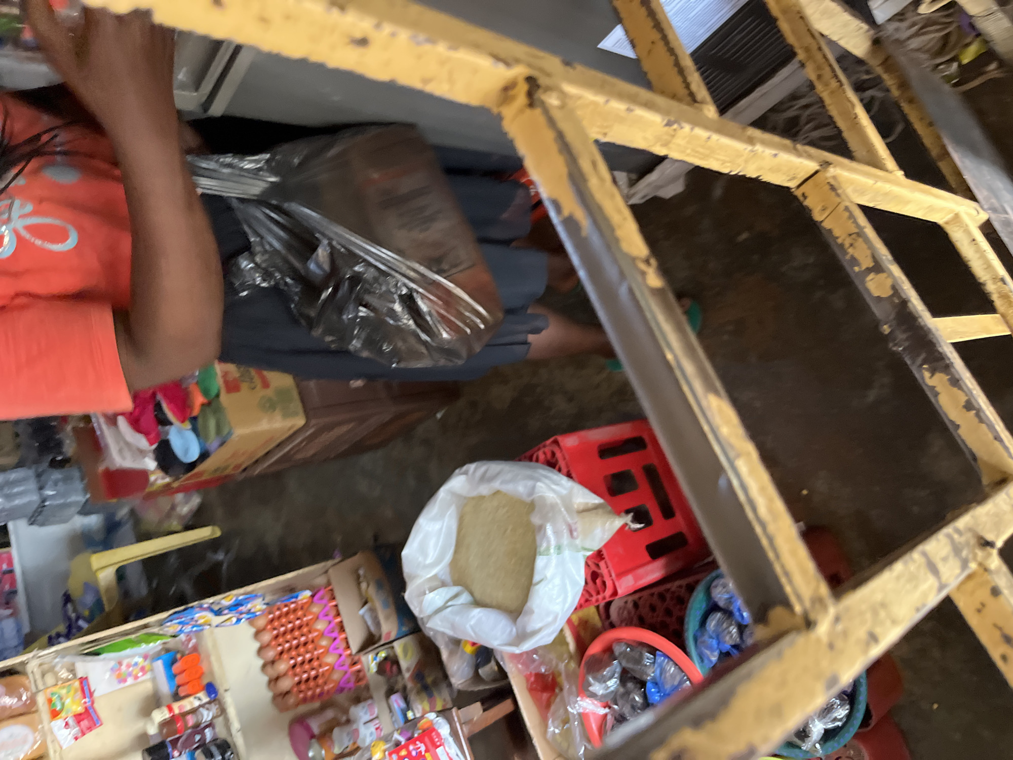 The image depicts a cluttered interior of what appears to be a small shop or market stall in Bukuru, Nigeria. In the foreground, a section of a yellow, metal ladder or shelving unit is visible, showing signs of wear and peeling paint. Behind the ladder, a person is partially visible, wearing an orange t-shirt with a light blue graphic and a dark skirt. They are holding a large, black plastic bag, which appears to be filled with items.
To the left of the person, a white plastic bag filled with what looks like rice or a similar grain is placed on a red plastic crate. Below this, a red plastic basin contains several small, dark, individually wrapped items, possibly snacks or candies. Further to the left, shelves are stocked with various goods, including boxes of eggs, bottles, and other packaged products. The floor appears to be concrete or dirt, with some debris visible. The overall impression is of a busy, functional retail space.