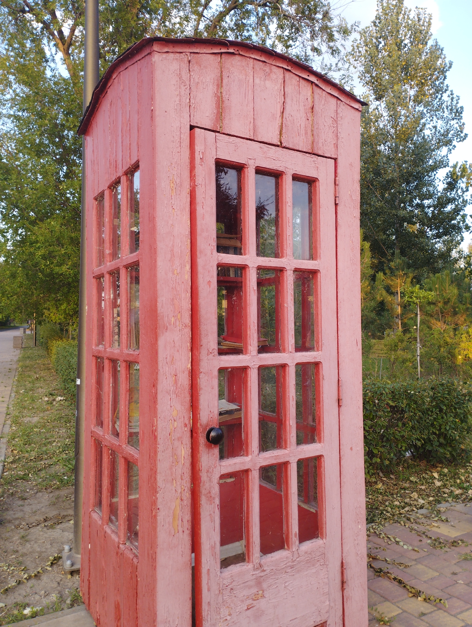 A weathered, red wooden booth with multiple glass panes stands outdoors. The booth appears to be an old phone booth, the paint peeling and the wood showing signs of age. It is positioned on a paved walkway next to a grassy area with fallen leaves. Trees and bushes form the backdrop, suggesting a park or public space. A tall, dark pole is visible to the left of the booth. This scene is located in the city of Rostov-on-Don, Russia.