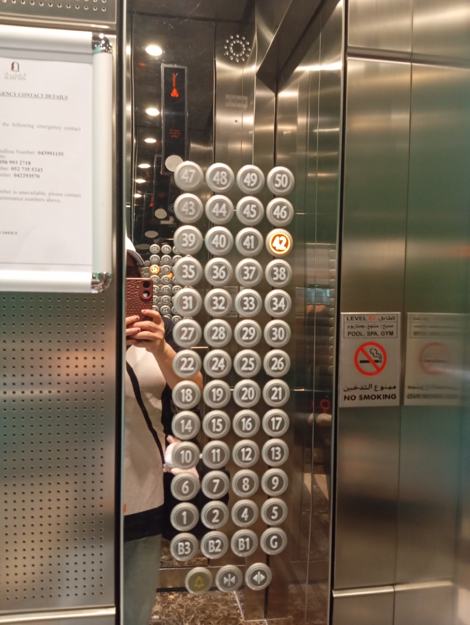 In Dubai, United Arab Emirates, a medium shot captures an interior view of a modern elevator. The elevator walls are highly reflective, showcasing a mirrored surface and brushed stainless steel. A person, partially visible in the reflection, is holding a pink smartphone and taking a selfie. The elevator control panel is prominently displayed, featuring numerous circular buttons for floor selection, ranging from numerical designations like "47" and "42" to alphanumeric labels like "B3" and "G." One button, marked "42," is illuminated, indicating the current or selected floor. To the right of the control panel, a sign in English and Arabic displays "LEVEL 02" and lists amenities such as "POOL, SPA, GYM," along with a "NO SMOKING" symbol. The elevator's interior is clean and well-lit, with visible ceiling lights and subtle reflections of the surrounding environment.