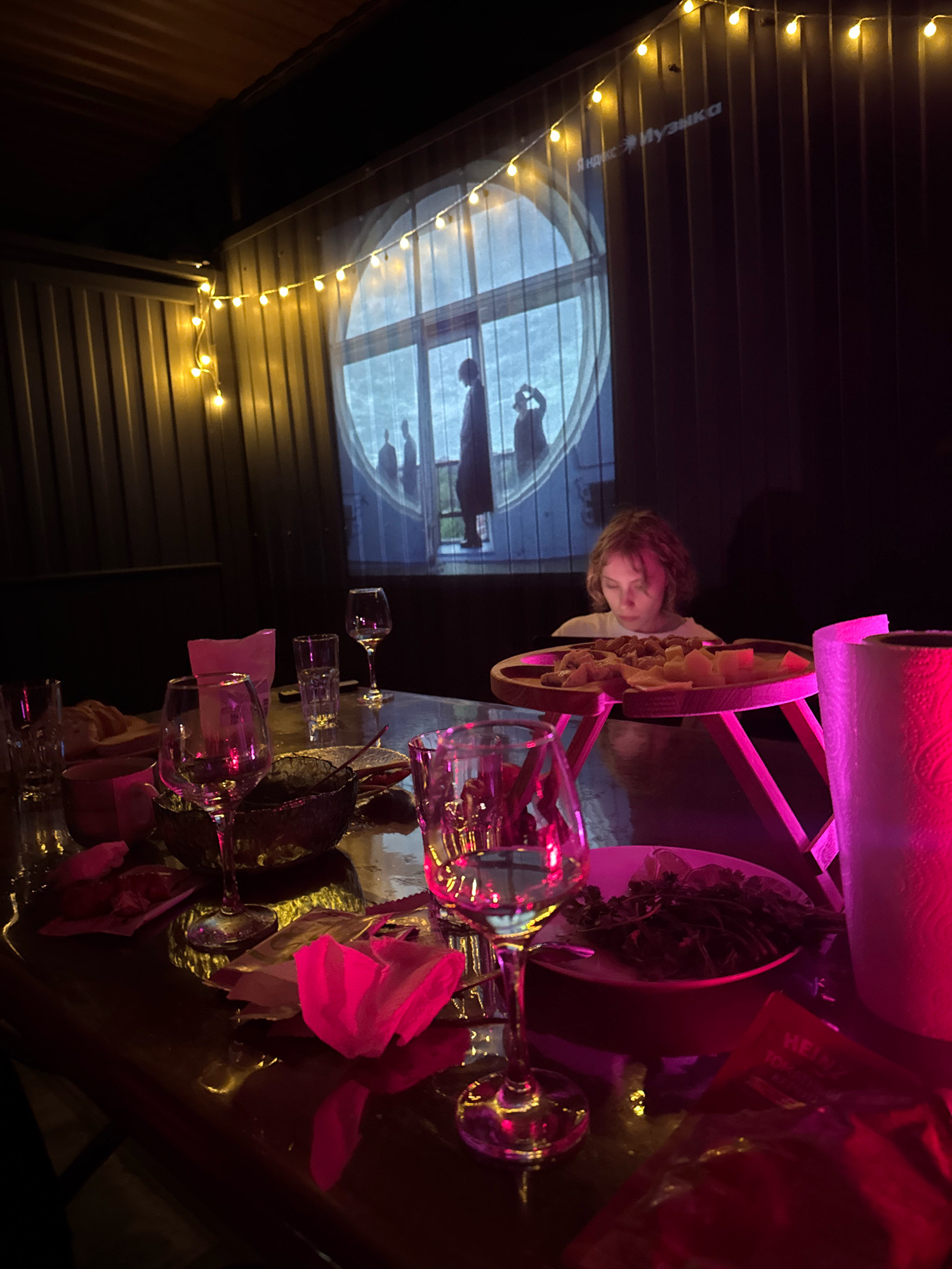 The image depicts a dimly lit outdoor seating area, possibly a patio or terrace, decorated with string lights. A young person with light brown hair is seated at a table, looking down at a wooden tray filled with food, likely an appetizer platter. The table is laden with wine glasses, plates of food, and what appear to be napkins, suggesting a meal or gathering. In the background, a projection is displayed on a corrugated metal wall, showing silhouettes of people within a circular frame, overlaid with Russian text that reads "Яндекс Музыка" (Yandex Music). The overall atmosphere is casual and convivial. The image does not contain any explicit information indicating its location in Tulskiy, Russia.