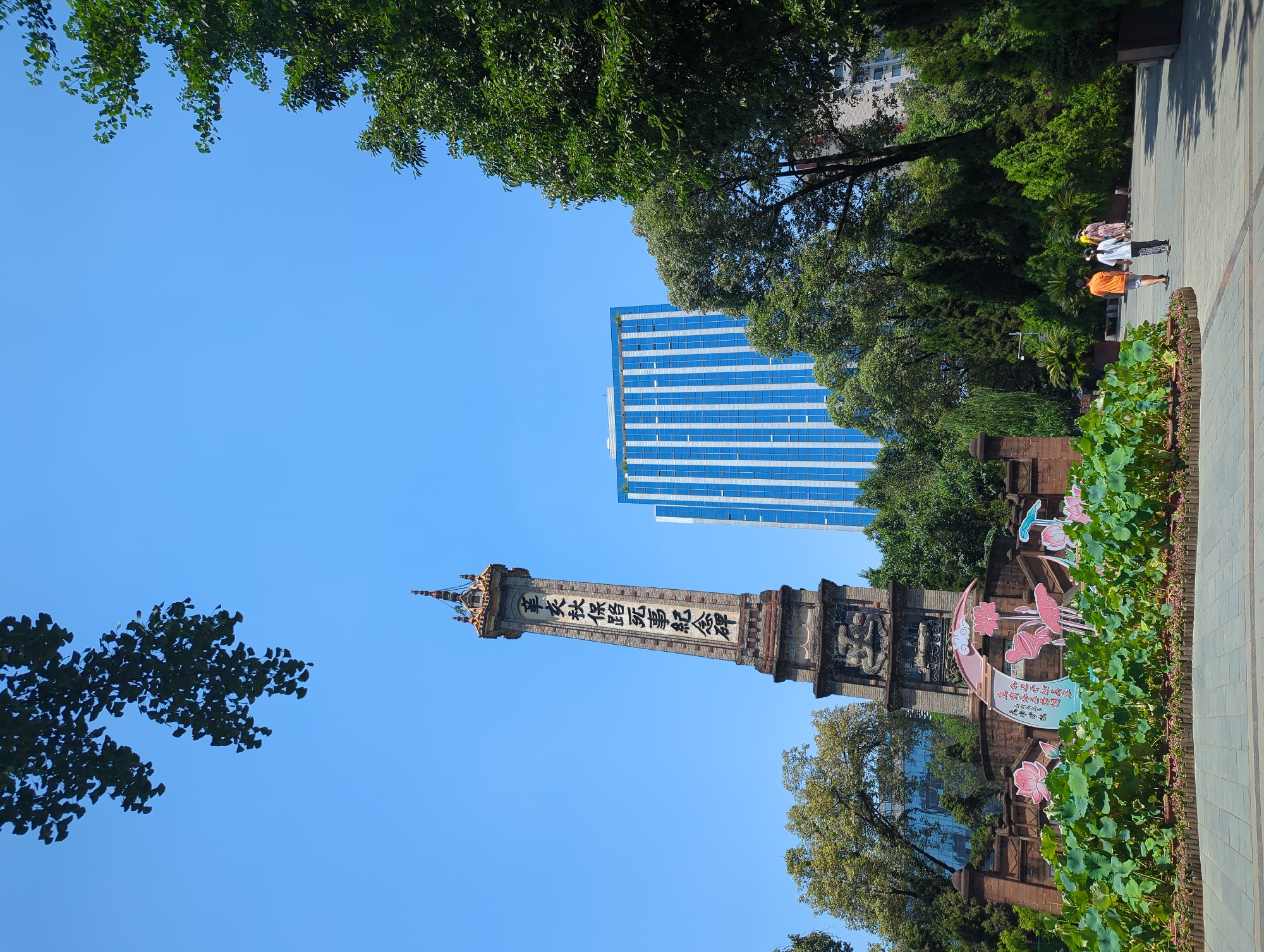 In the city of Qingyang District, China, a tall, weathered monument with Chinese characters inscribed vertically on its side stands under a clear blue sky. The monument appears to be made of stone and has intricate carvings at its base. In front of the monument, a colorful decorative display featuring stylized pink lotus flowers and a crescent moon shape is visible, surrounded by lush green plants. To the right of the monument, a modern, tall building with a blue facade and white vertical stripes dominates the background. Several trees with dense green foliage are scattered throughout the scene, with some branches reaching into the upper portion of the frame. A paved pathway runs through the foreground, and three individuals are walking away from the camera on this path. The environment suggests a public park or outdoor recreational area.