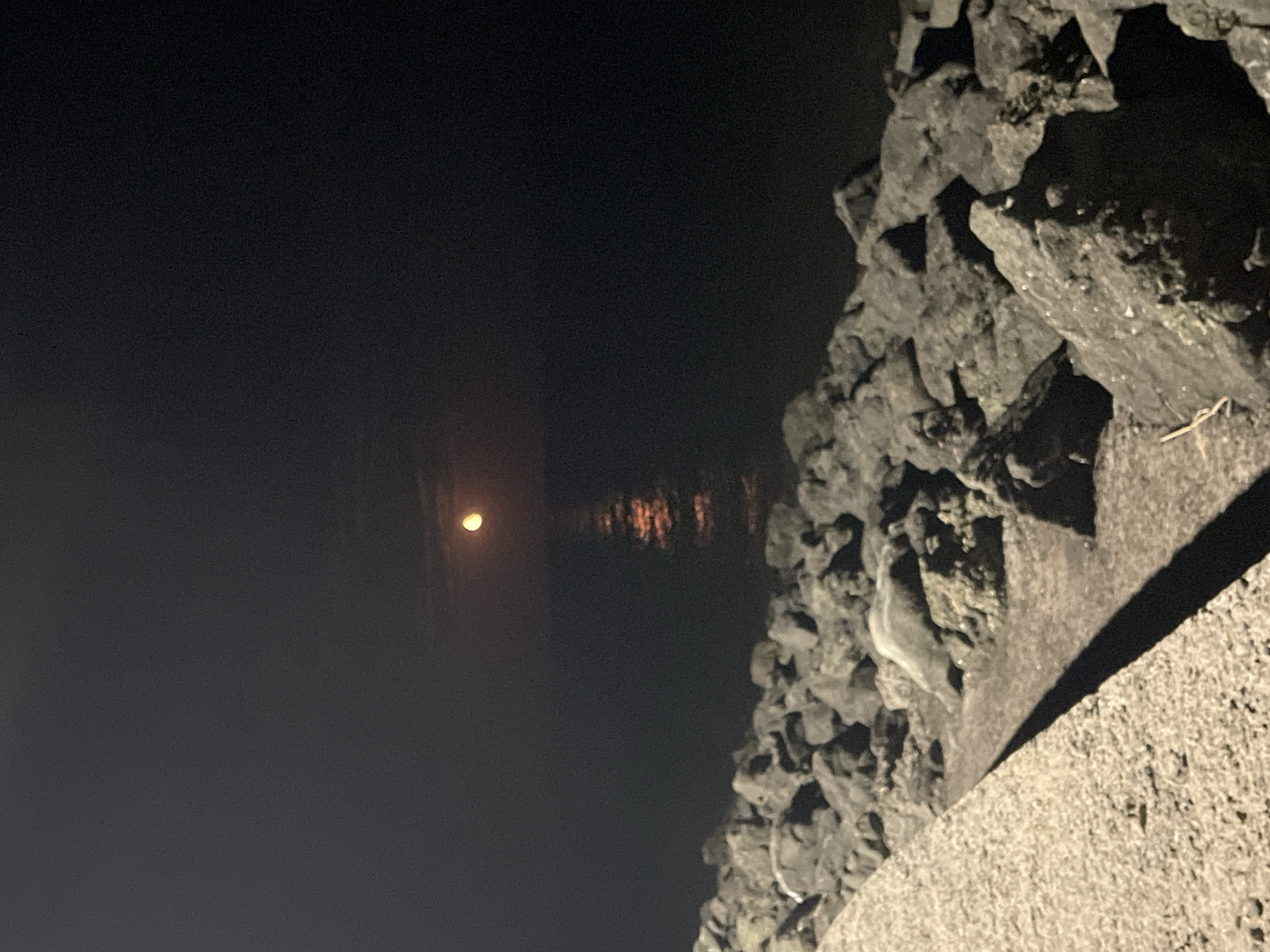 The image depicts a nocturnal scene at a coastal area, likely in Primorsko, Bulgaria. In the upper half of the image, a luminous, orange-hued moon hangs in a dark, overcast sky, its reflection shimmering on the calm surface of the sea below. The sea extends towards the horizon, which is barely discernible due to the darkness. The lower half of the image features a rocky shoreline. Large, dark-colored rocks, some appearing wet, are piled along the edge of the water. In the foreground, a concrete embankment or walkway runs diagonally from the bottom left corner upwards, casting a long shadow. The overall impression is one of a quiet, somewhat stark, natural landscape illuminated by the moon.