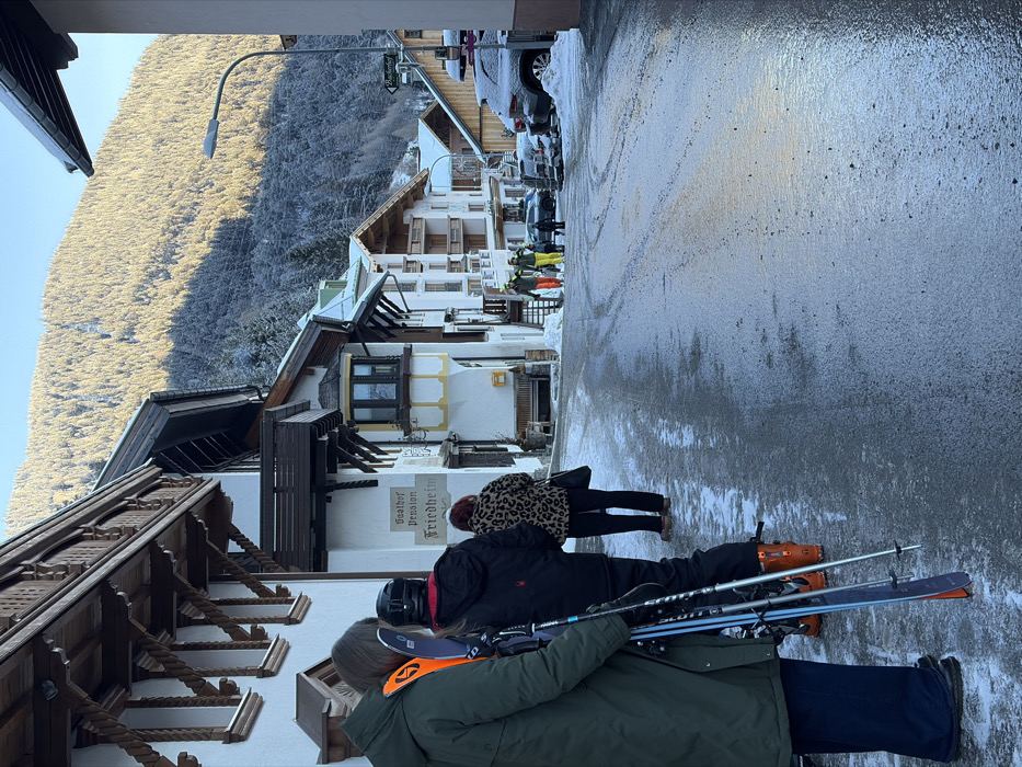 A street view in Sankt Jakob am Arlberg, Austria, captures a winter scene under clear skies, with the ground partially covered in snow and ice. The asphalt road in the foreground displays wet patches and tire tracks. On the left side of the road, three individuals are positioned with their backs to the viewer. The leftmost person wears a dark green winter coat and holds a pair of orange and black skis with attached poles. The adjacent person wears a black ski jacket, a black helmet, and orange ski boots, also holding ski poles. The third individual wears a leopard-print winter coat and dark pants, carrying a dark shoulder bag.
Further down the street, a group of at least three, possibly four, individuals, including children, walk away from the camera, dressed in bright winter attire, including helmets and colored jackets (yellow, green). Traditional Alpine buildings line both sides of the street, featuring multi-story structures with wooden balconies, decorative supports, and snow-covered roofs. A prominent sign on a white building to the left reads "Gasthof Pension Friedheim." A wooden bench is visible outside this building. On the right, further down the street, several vehicles, including SUVs, are parked and partially covered in snow. A streetlamp stands on the right side. The background is dominated by a snow-covered, forested mountain, with a clear shadow line indicating direct sunlight on the upper slopes.