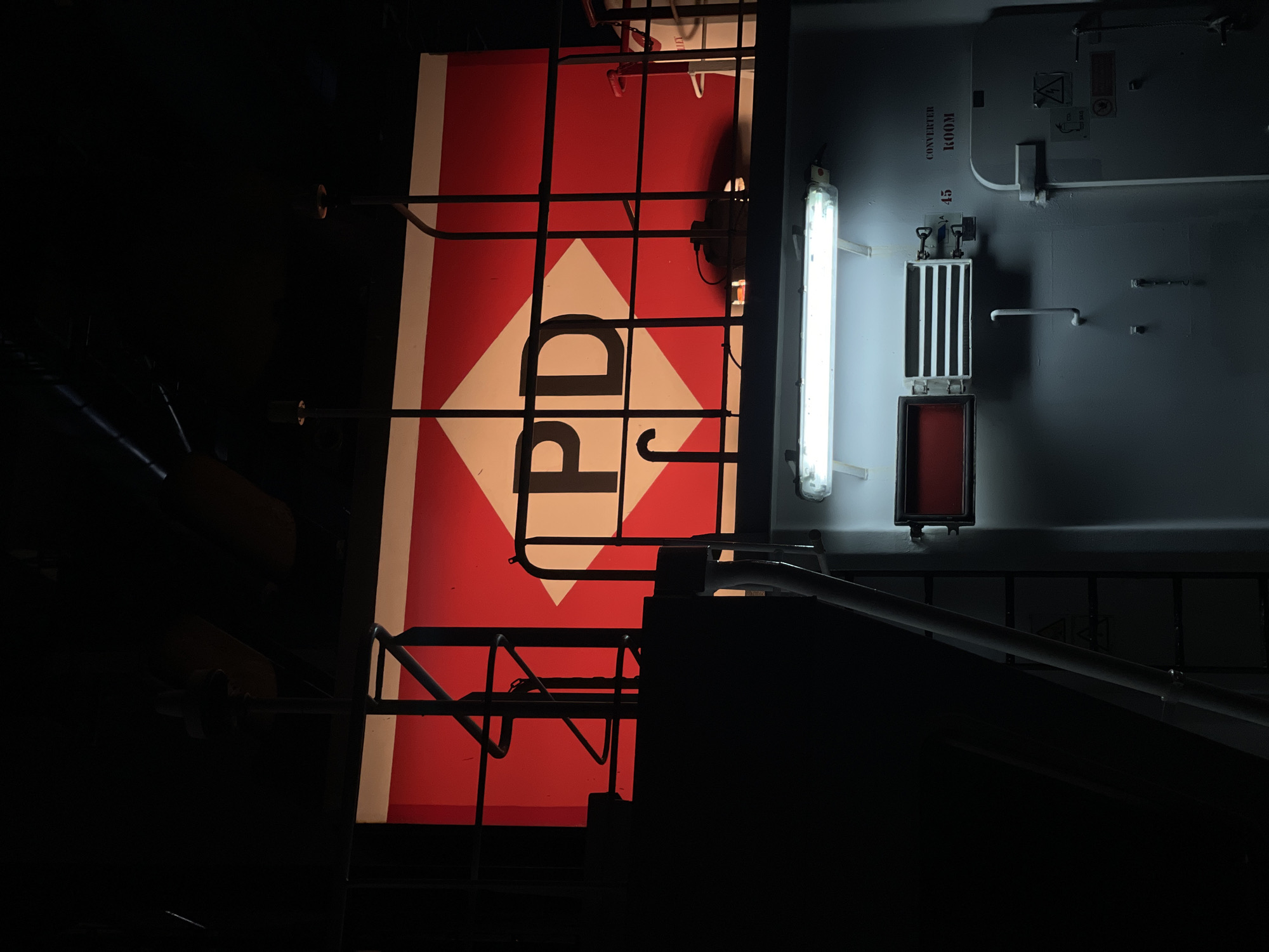The image depicts an industrial or maritime setting, possibly a ship or a port facility, in Abidjan, Ivory Coast. Dominating the upper portion of the frame is a large, illuminated red and white diamond-shaped sign with the letters "PD" prominently displayed in black. This sign appears to be mounted on a structure with railings and a staircase, suggesting an elevated walkway or platform.

Below this sign, the environment transitions to a more enclosed space, illuminated by fluorescent lighting. A light fixture runs horizontally across the wall. To the left of the fixture, a rectangular red indicator is visible, and to its right, a metallic panel with horizontal slats is mounted. Further to the right, text stenciled onto the wall reads "CONVERTER ROOM" and the number "45". A large, off-white door with latches and warning symbols is also present. The overall atmosphere is dim and utilitarian, with stark lighting and industrial features. There are no people visible in the image. The scene is captured from a slightly upward angle, emphasizing the scale of the signage and the structure above.