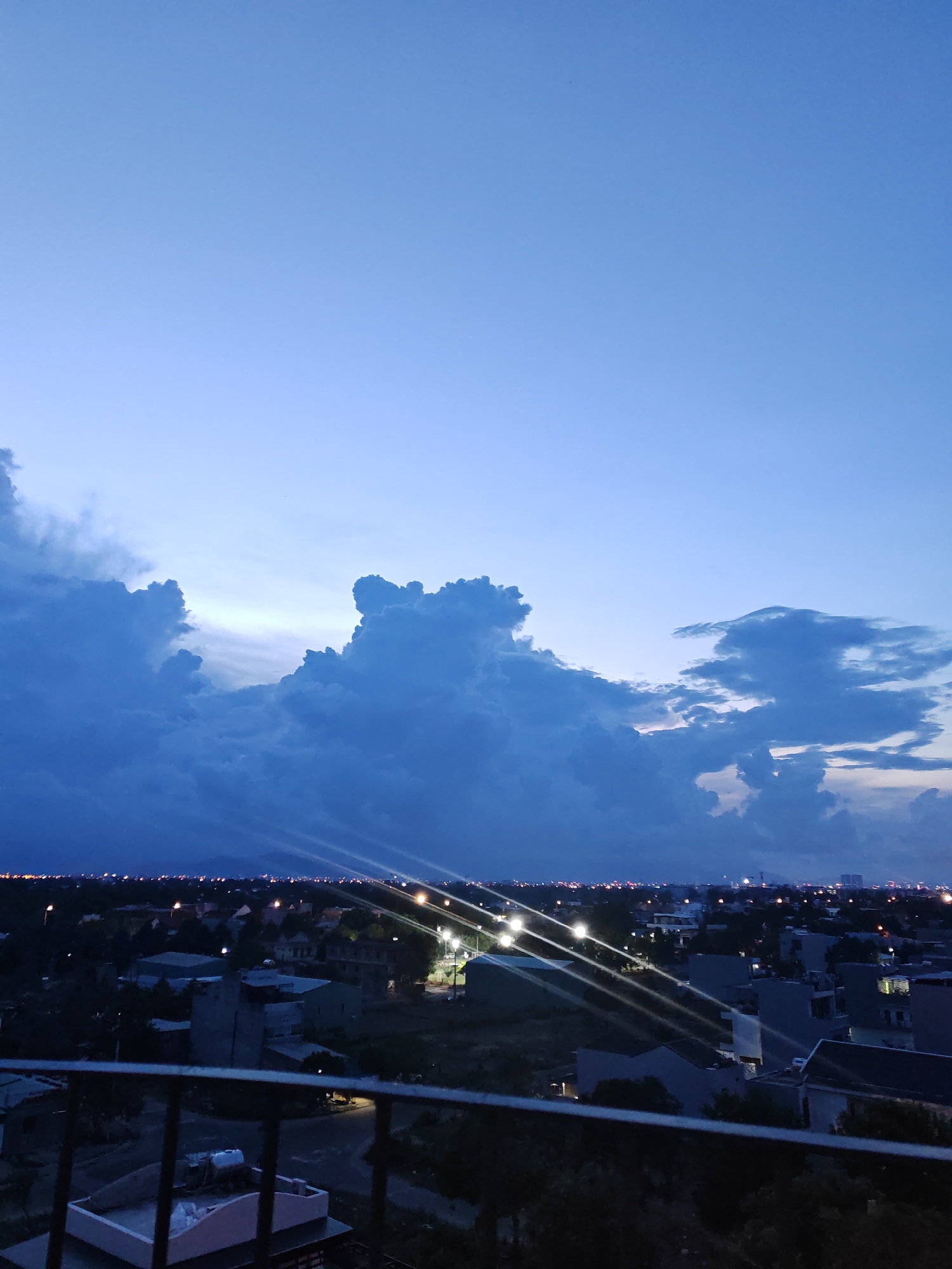 The image depicts an aerial view of a city at dusk, likely Đà Nẵng, Vietnam, with a broad expanse of the sky dominating the frame. The sky transitions from a deep, almost navy blue at the top to lighter hues of lavender and pale blue near the horizon, suggesting the last moments of twilight. Dramatic, dark blue clouds, heavily textured and contoured, are clustered in the middle of the sky, with some forming towering, mountainous shapes. A softer, brighter band of light is visible behind these clouds, hinting at the fading sunlight.
Below the sky, the cityscape is spread out, appearing as a dense collection of buildings and structures illuminated by artificial lights. Streetlights and lights from within buildings create a scattering of yellow and white dots across the urban landscape, outlining roads and structures. The buildings are a mix of low-rise and multi-story constructions, with some appearing to be houses and others more commercial or industrial in nature. The vegetation is dark and indistinct due to the low light conditions. In the foreground, a dark metal railing or balcony structure is visible, suggesting the vantage point is from a higher elevation, possibly a building or a bridge overlooking the city. The overall mood is serene and atmospheric, characteristic of a city winding down for the night.