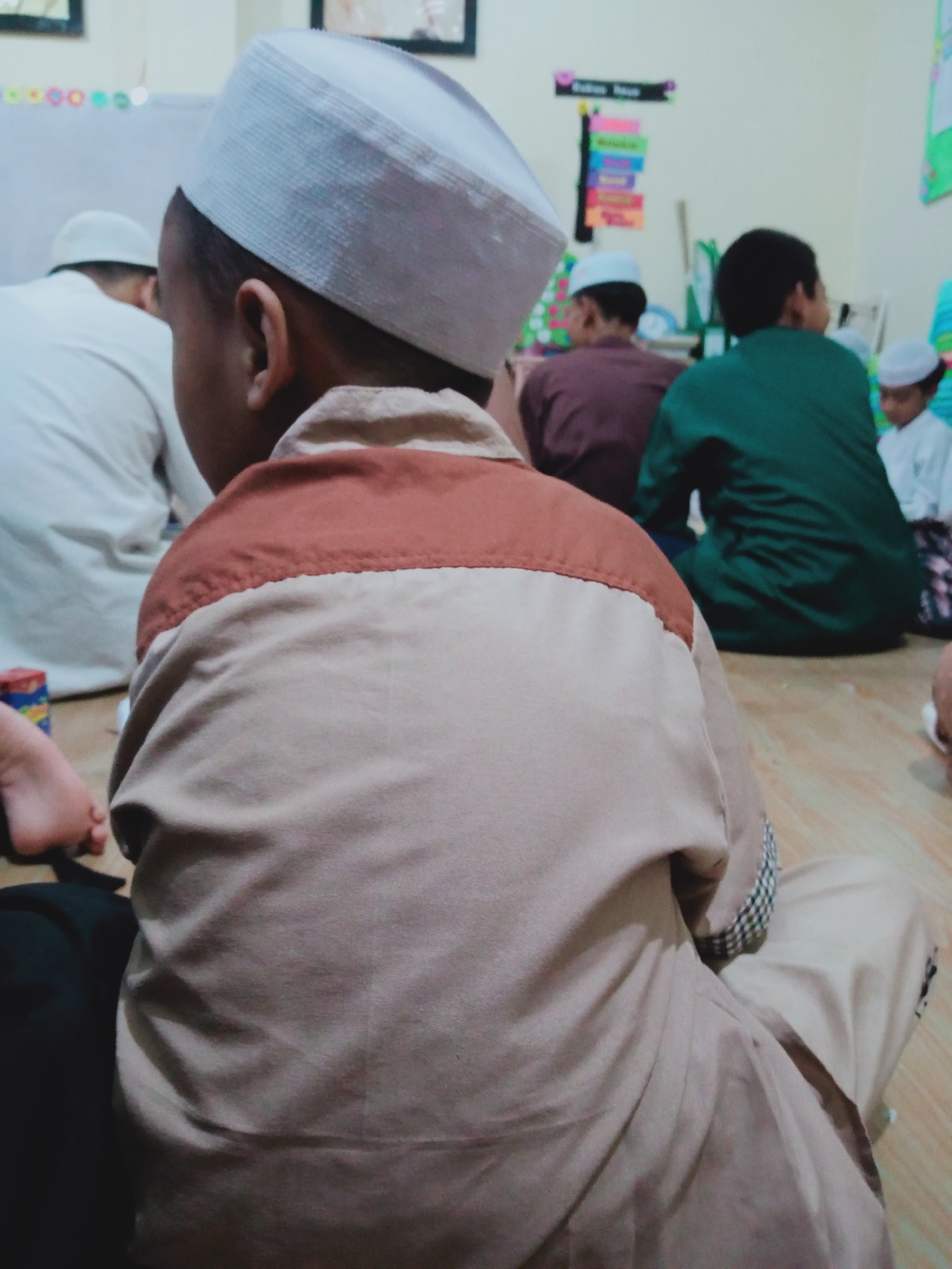 The image depicts a group of young boys seated on the floor, likely in a religious educational setting, such as a madrasah or recitation class. The primary subject is a boy in the foreground, facing away from the camera, wearing a light brown garment with a darker brown yoke and a white, checkered trim around the cuff. He is also wearing a white peci, a traditional cap. Behind him, several other boys are visible, also wearing pecis and similar modest clothing. One boy in the middle ground wears a dark maroon garment, and another to his right wears a bright green one. Further back, a boy in a white peci and garment is partially visible. The environment appears to be indoors, with a wooden floor and a light-colored wall in the background. Educational posters with colorful lettering are affixed to the wall. The overall impression is one of a group engaged in a learning activity, common in East Jakarta, Indonesia.