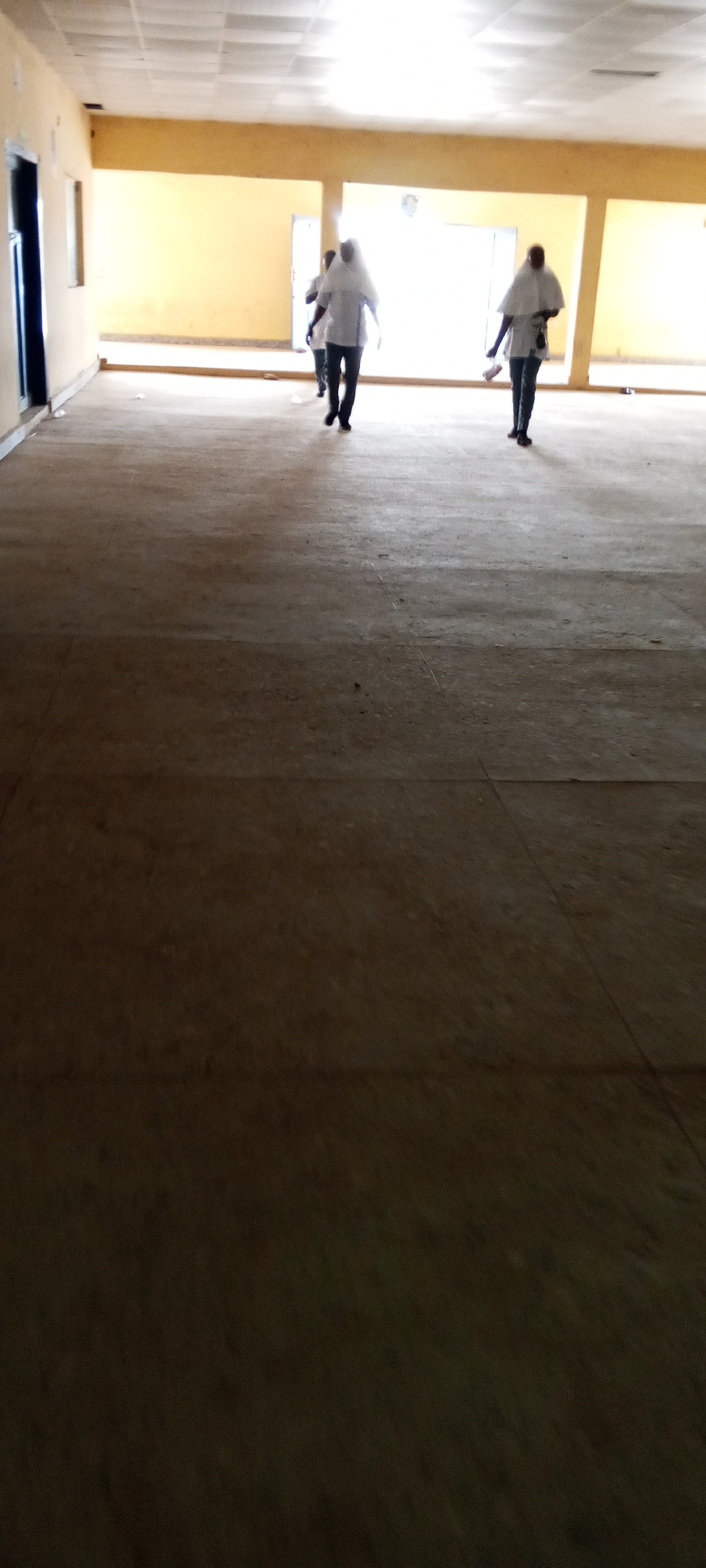 The image depicts an interior space in Unknown, Unknown, possibly a large, sparsely furnished room or hall. The floor is primarily a dark, mottled brown with visible seams suggesting tiling or large concrete slabs. The walls are painted a pale yellow or beige, with diffused natural light entering from several large windows or doors along the far wall, creating a brightly lit zone. Three people are walking towards the camera in the distance. They appear to be wearing white shirts or lab coats and dark pants. Their faces are obscured due to distance and lighting conditions. The person on the right seems to be holding something. A door is visible on the left side of the image. The scene appears to be in a state of transition or preparation.
