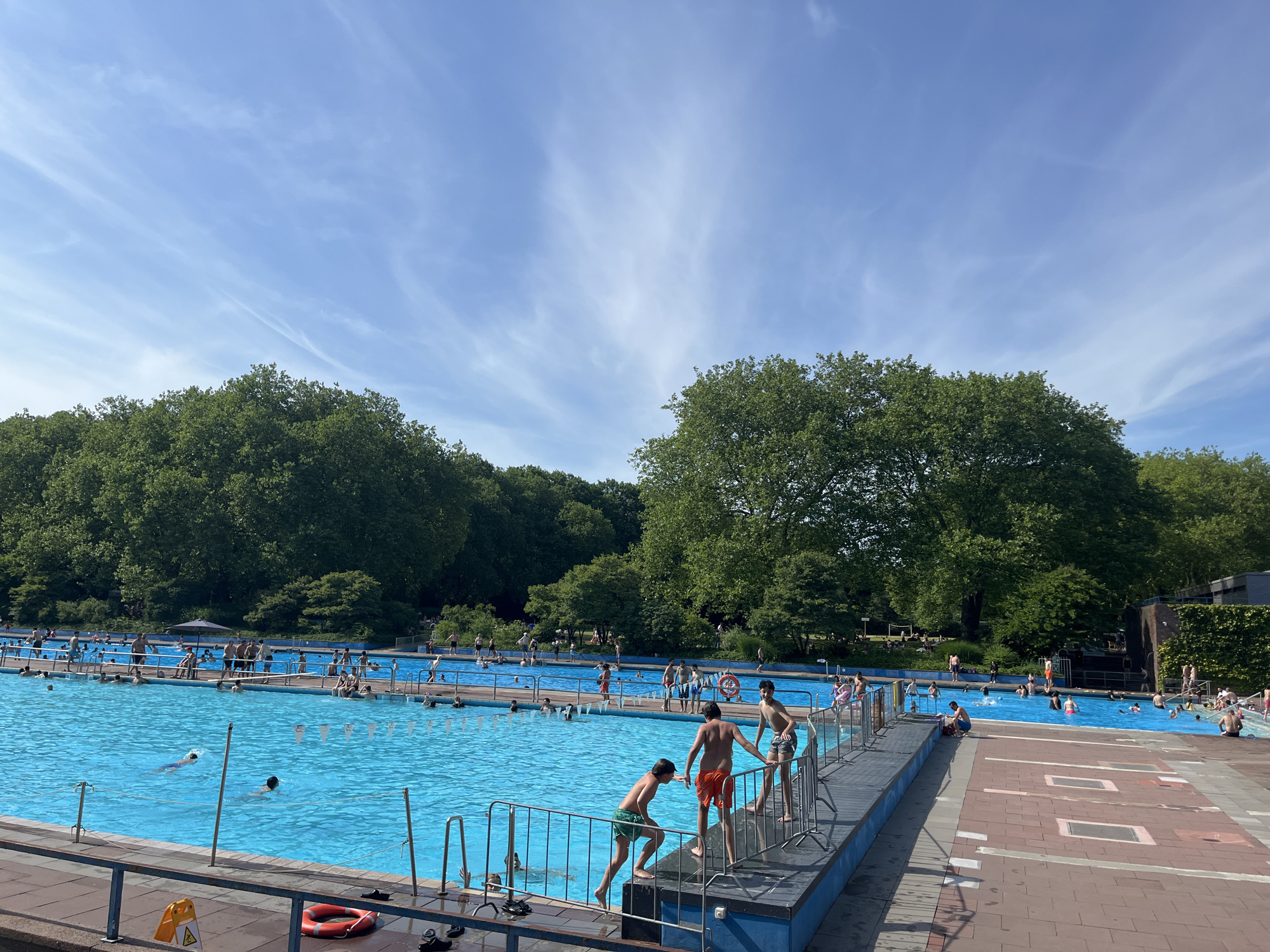 This is a daytime, eye-level, wide shot of a crowded outdoor swimming pool complex in Gladbeck, Germany. The pool is a bright, clear blue with numerous swimmers visible, some actively swimming and others wading or relaxing. Along the edge of the pool are tiled walkways and pathways, interspersed with brick-like patterns. A metal fence separates the pool area from the viewer.
In the foreground, on the right side of the image, three young males are visible on a raised platform. One boy, wearing green swim trunks, is stepping over the railing into the pool. Another boy, in orange swim trunks, is standing next to him, looking down. A third boy, wearing checkered swim trunks, is standing further back. On the ground near the railing, a red life preserver and some black footwear are visible.
The background of the image is dominated by lush green trees, indicating a park-like setting surrounding the pool. The sky is a bright blue with wispy white clouds. Other swimmers and people are visible in the distance, scattered throughout the pool and on the surrounding decks. Some are under umbrellas, suggesting shaded areas.
The overall impression is a lively, recreational scene on a sunny day.