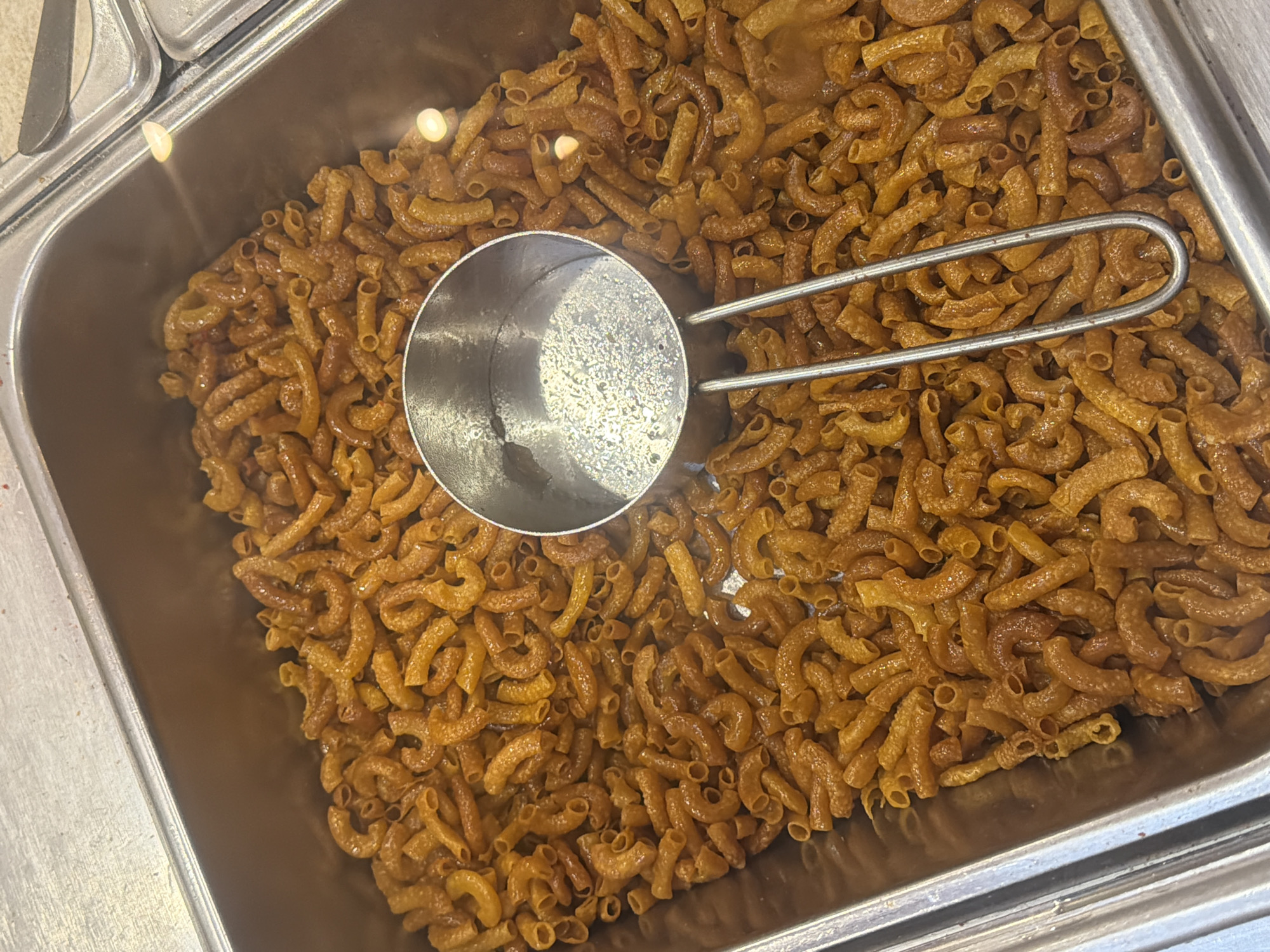 The image shows a close-up, overhead view of a metal tray filled with macaroni. A metal scoop with a curved handle rests on top of the pasta. The macaroni pieces are curved and appear to be coated in a sauce or glaze, giving them a shiny, golden-brown appearance. The tray itself is part of a larger serving setup, with another section visible to the left. The lighting suggests an indoor setting, possibly a food service establishment. The scene is still and focused on the food. No people, text, time of day, or weather details are discernible. The location context is East Jakarta, Indonesia.