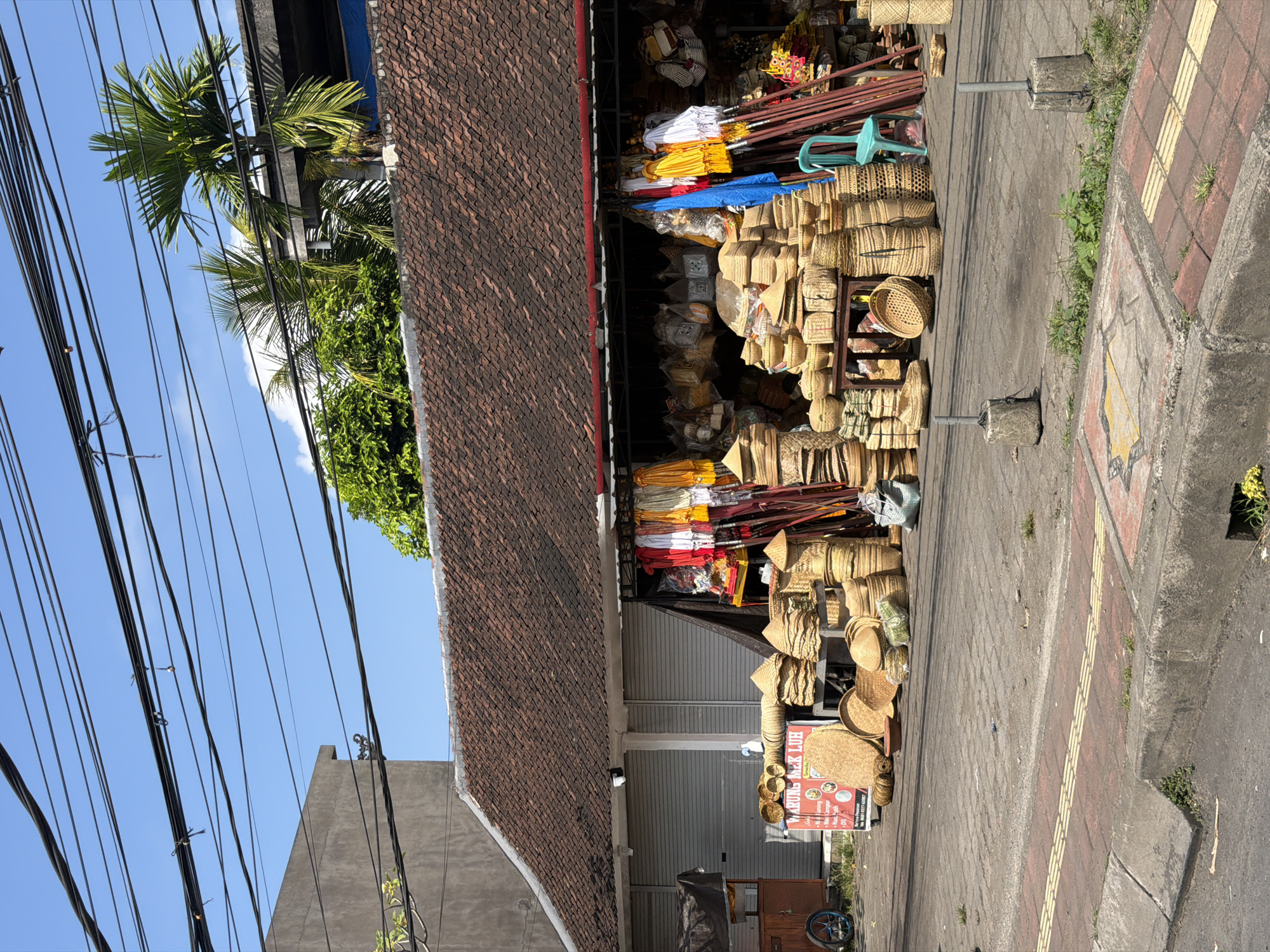 The image depicts a street-side market stall in Blahbatuh, Indonesia, filled with a variety of woven goods. The stall has a traditional tiled roof and is open to the street, displaying numerous woven baskets, hats, mats, and bags. In front of the stall, the items are arranged on the pavement and on low shelves. Some of the woven items are conical hats, while others are larger, round baskets. Behind the display of woven goods, there are colorful fabrics and umbrellas hanging, as well as more woven bags and other products stored within the stall. A bright blue plastic chair is visible to the right of the display. The street in front of the stall is paved with bricks and concrete, with a curb and a drainage grate. A sign with Indonesian text is propped up near the front of the stall. In the background, there are closed metal roller doors of a building, palm trees, and overhead electrical wires. The sky is clear and blue. There are no people visible in the image.