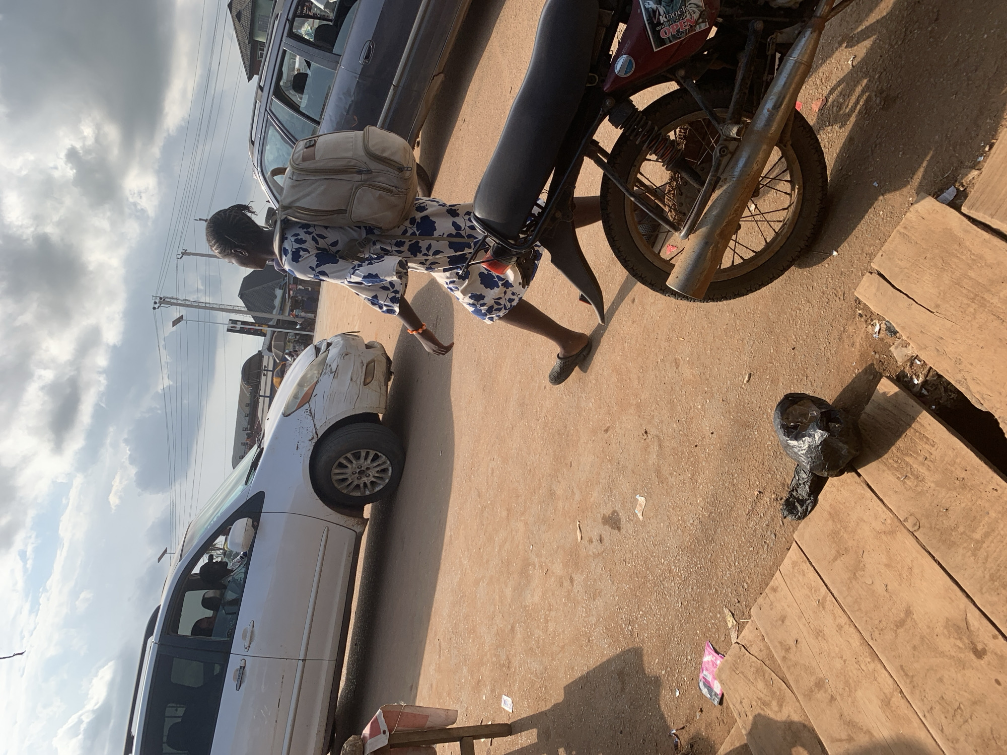 A young woman with braided hair wearing a blue and white floral dress and carrying a light-colored backpack walks along a dusty road. She is wearing dark, flat shoes. Beside her, a motorcycle is parked, its red body visible. The motorcycle has a sticker on its side that reads "Knock on the door...Open." Further down the road, a white minivan is stopped, with a driver visible inside. Behind the minivan, another dark-colored vehicle is partially visible. The scene appears to be a street in Shagamu, Nigeria, under a cloudy sky. The sunlight casts long shadows, suggesting it is either morning or late afternoon. The ground is covered in reddish-brown dirt, indicative of a dry season. Scattered on the ground are pieces of trash, including a black plastic bag. The overall atmosphere suggests a busy, perhaps traffic-laden, urban or semi-urban environment.