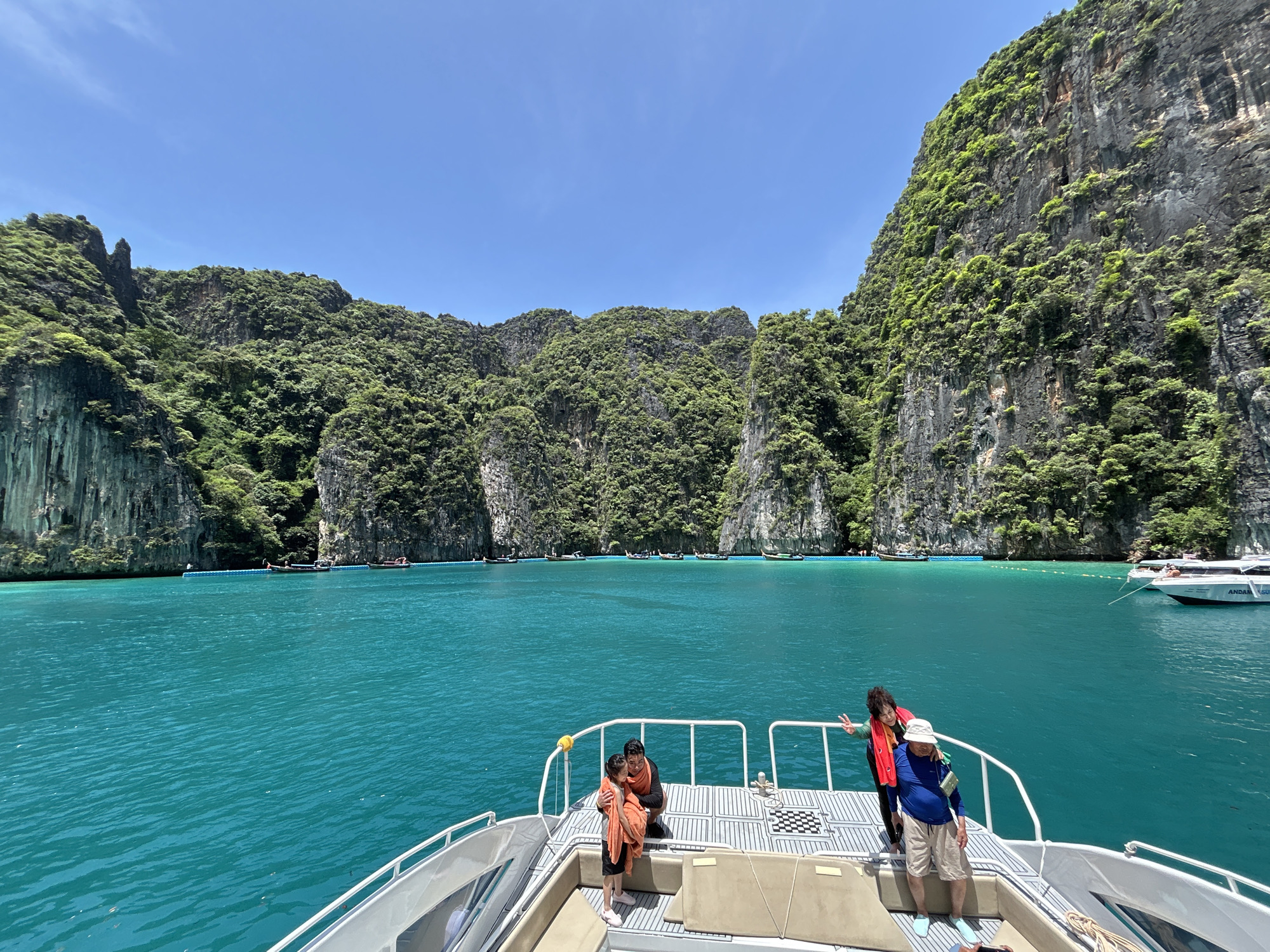 In Ao Nang, Thailand, on a sunny day, a speed boat navigates through incredibly clear turquoise waters. Towering, lush green limestone cliffs rise dramatically from the water, creating a breathtaking natural amphitheater. Several long-tail boats and a larger white speed boat are moored or anchored in the bay. On the deck of the foreground speed boat, three people are visible. A man in a blue shirt and beige shorts, wearing a white hat, stands with his arm around an older woman in a red life vest who is giving a peace sign. Nearby, a man in a red shirt and a young girl in an orange wrap are embracing. The boat's white railing and seating are also visible.
