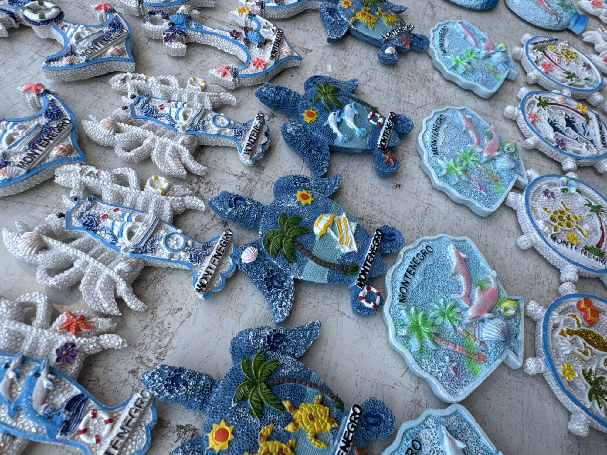 The image captures a close-up, overhead view of a diverse collection of souvenir items, likely magnets, scattered across a light, textured surface resembling concrete or stone. The bright, natural lighting suggests an outdoor setting during daytime.
The subjects are exclusively these decorative objects, which are themed around marine life and coastal motifs, indicative of a tourist destination such as Budva, Montenegro. The collection features a variety of shapes:
* **Blue turtles:** Several blue-colored turtles are depicted, adorned with details like palm trees, yellow suns, white dolphins, miniature yellow turtles, and life preservers. Many of these have a glittery texture.
* **White starfish/crab-like shapes:** Numerous white, textured items resemble starfish or crabs, decorated with blue maritime patterns, small red starfish, purple flowers, and miniature shells.
* **Country outline shapes:** Some light blue and white pieces are shaped like an outline of a country, featuring internal scenes with pink dolphins, palm trees, and shells.
* **Boat shapes:** Other items are shaped like small boats, typically in white and blue.
A highly prominent detail across nearly all visible souvenirs is the word "MONTENEGRO", clearly identifying the items as mementos from the country. The predominant colors are various shades of blue and white, accented with vibrant pops of yellow, red, green, and pink in the decorative elements. The items appear to be mass-produced tourist merchandise. No human activity or specific event is taking place; the scene is a still life of these commercial products.