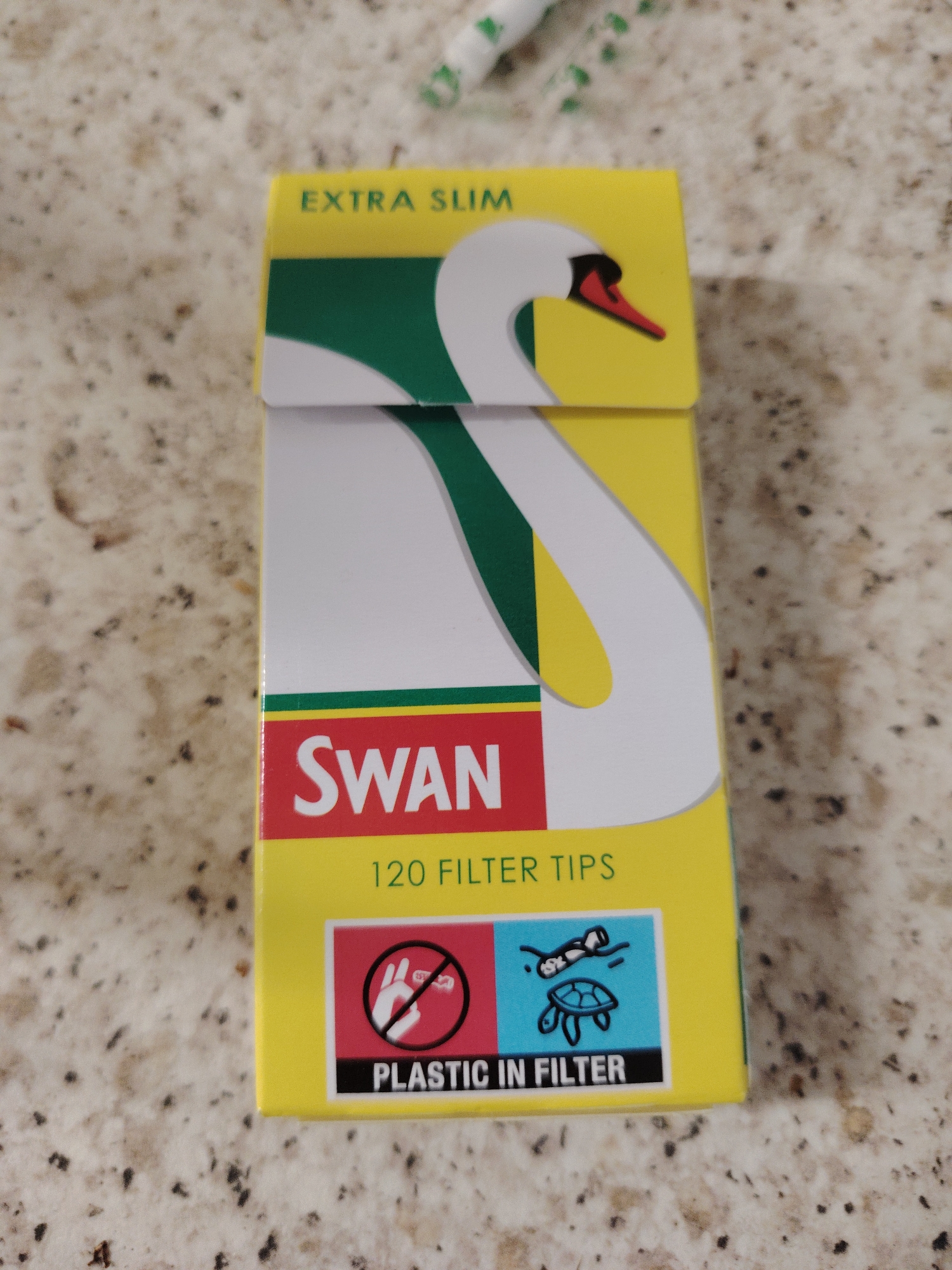 The image displays a brightly colored yellow and green box of "Swan" brand filter tips, centrally placed on a speckled, light-brown countertop. The box's design prominently features a stylized white swan with a red beak, its form curving across the front and top panels against a green and yellow background.
Visible text on the box includes "EXTRA SLIM" at the top, "SWAN" in large red letters on a white band below the swan's neck, and "120 FILTER TIPS" beneath it. At the bottom of the box, a black stripe carries the bold warning "PLASTIC IN FILTER". Flanking this text are two small informational symbols: on the left, a white hand is shown dropping a filter tip into a trash can, encircled by a red "no" symbol, indicating "do not litter"; on the right, a blue square depicts a turtle with small, dark shapes floating above it, symbolizing marine life affected by plastic pollution.
The background is an out-of-focus, light-colored surface with dark speckles, likely a kitchen counter or table. A blurred, white and green object, possibly a rolled item or another filter, is partially visible in the upper right background. The scene is a still life, with no people or specific activities taking place, and appears to be an indoor setting under ambient light in Huntingdonshire, United Kingdom.