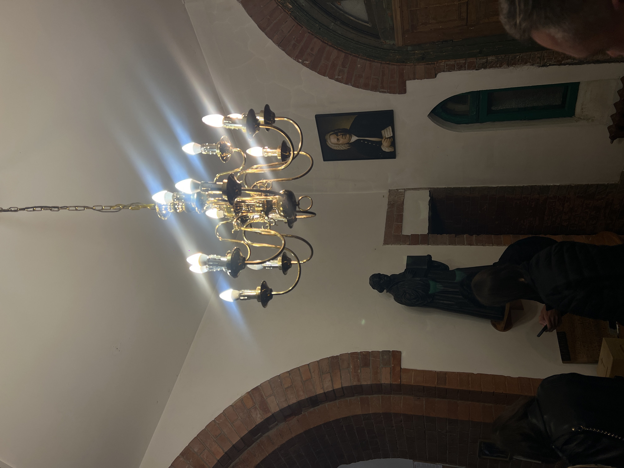 The image captures an interior view of the Lutheran Church of St. Paul in Vladivostok, Russia. A large, ornate golden chandelier with ten illuminated candelabra-style bulbs hangs centrally from the white ceiling, casting bright light and lens flares. Below the chandelier, the scene reveals white walls accented by prominent red brick arches on both the left and right sides, indicating a historical architectural style.
On the left, a dark, robed statue, possibly depicting a religious figure holding a book, is mounted on a shelf. Below and to the left of the statue, the backs of two individuals with dark hair are visible, one wearing a black leather jacket. A third person, on the right foreground, has only the back of their head and shoulder visible. Another individual in the mid-ground is partially visible from behind, looking down at a smartphone.
To the right of the statue and under the chandelier, a framed portrait of Johann Sebastian Bach is affixed to the wall, depicting him with a white wig, dark coat, and holding a piece of paper. Further to the right, a tall, narrow window with a dark green arched frame is set into the wall, flanked by brickwork. The lower section of the window appears opaque or frosted. The overall environment suggests a cultural or religious venue, possibly an event or concert.