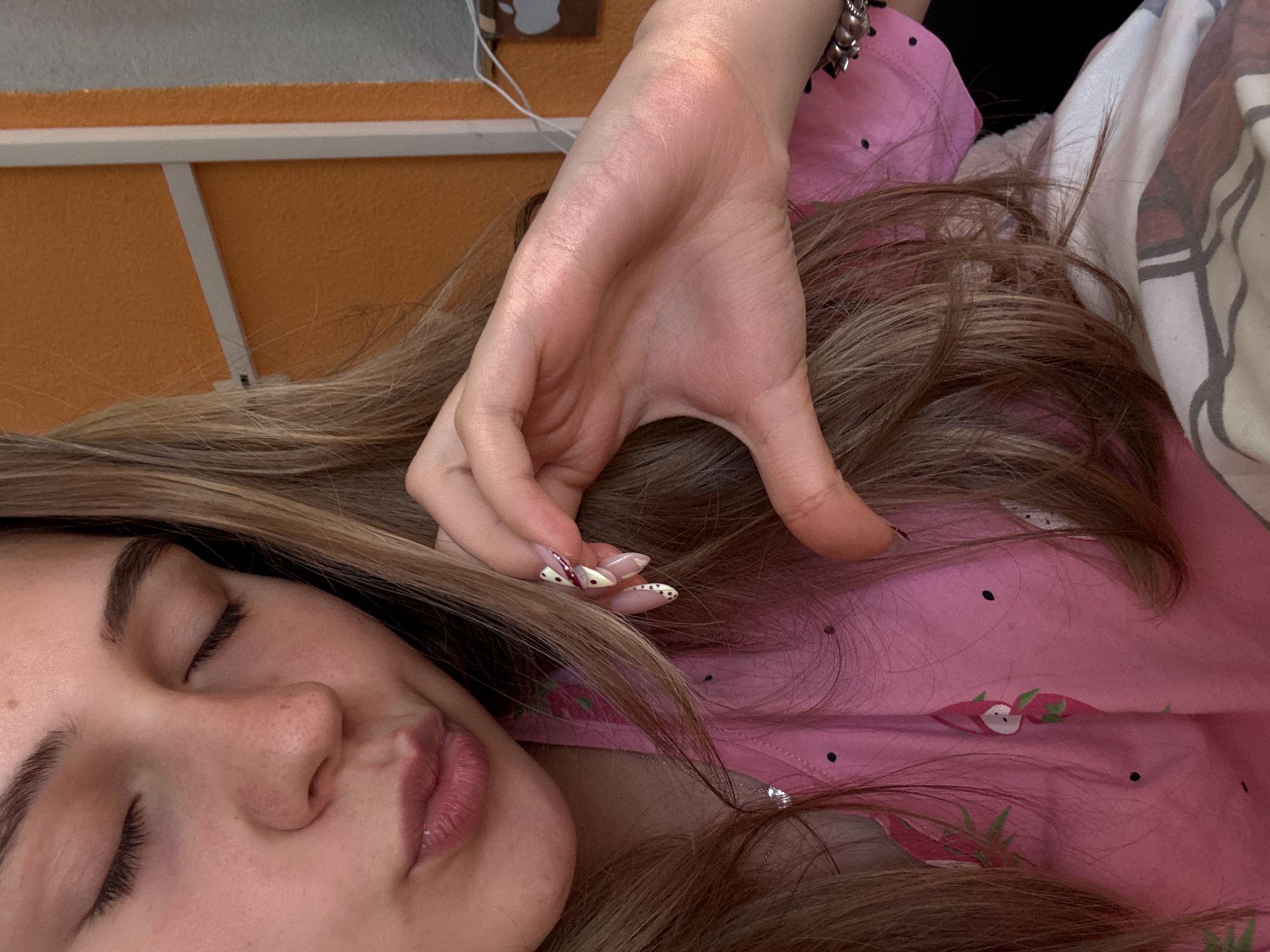 A woman lies on her back, eyes closed, with her right hand resting on her light brown hair. She is wearing a pink floral-patterned shirt. The background shows a portion of an orange wall and a white shelf or frame. A wrist with a bracelet is visible near the top right of the frame. There's also a white and patterned sheet. The location in the city of Panagyurishte, Bulgaria is not deducible from the image.