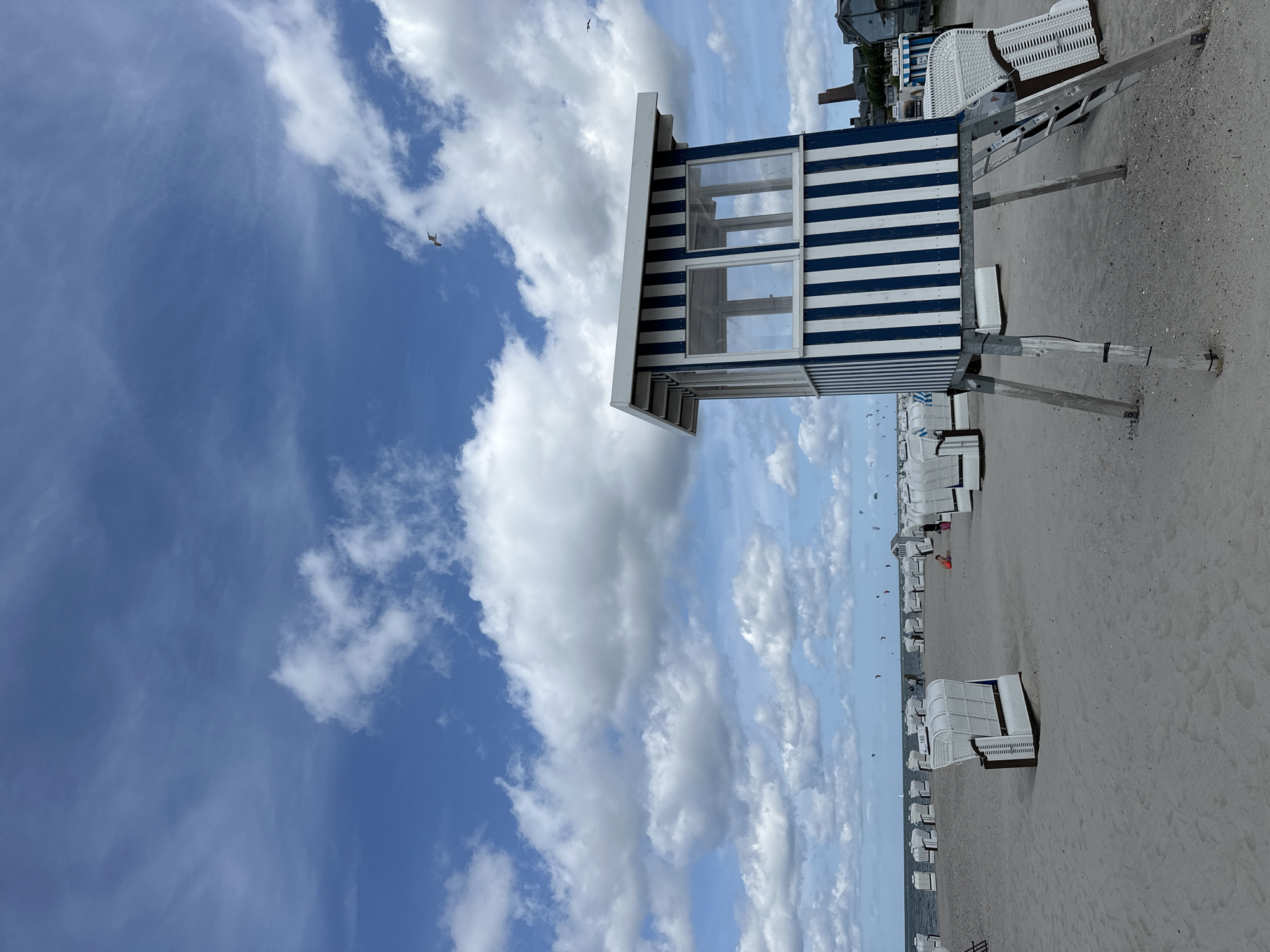 This is a photograph of the beach in Laboe, Germany, on a partly cloudy day. The foreground is dominated by a sandy beach with numerous white, wicker beach chairs. A blue and white striped lifeguard station, elevated on stilts, stands prominently on the right side of the frame. A ladder provides access to the station's elevated platform. In the background, the sea meets the horizon under a sky filled with puffy white clouds. Several kite surfers are visible on the water, and a few buildings can be seen in the distance on the right. Two seagulls are also in the sky.