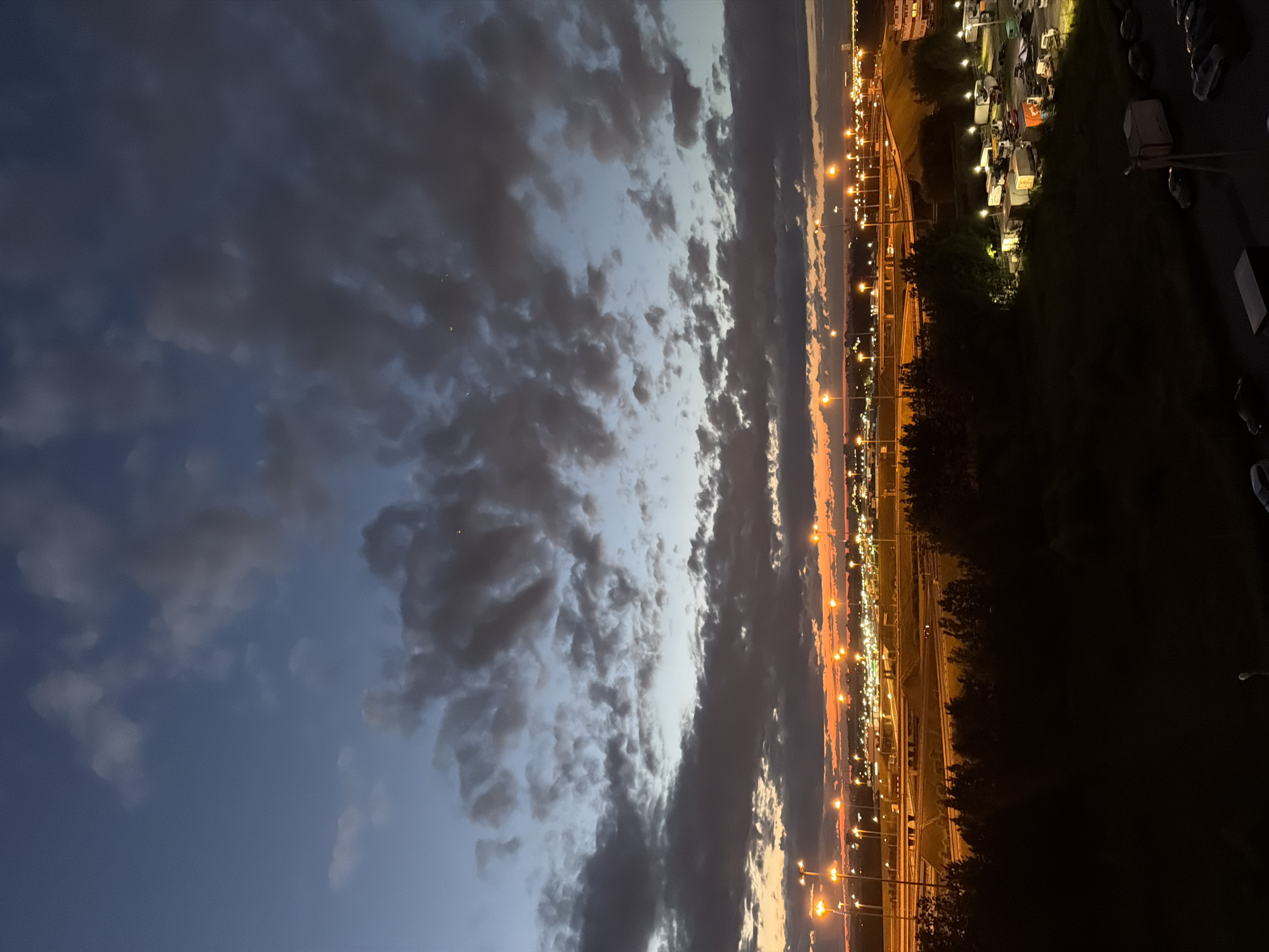 This is a vertical, eye-level, medium shot photograph taken at dusk from an elevated vantage point overlooking a city landscape. The sky is a dramatic gradient of deep blue, fading into lighter blues and oranges near the horizon, creating a sunset effect. Dark, textured clouds are scattered across the sky. Below the sky, a city sprawl is visible, illuminated by numerous streetlights and building lights. A complex network of elevated highways with bright lights stretches across the midground. In the foreground, a dark silhouette of trees partially obscures a parking lot filled with parked vehicles, including several trucks and cars. The scene conveys a sense of urban twilight and the transition from day to night.